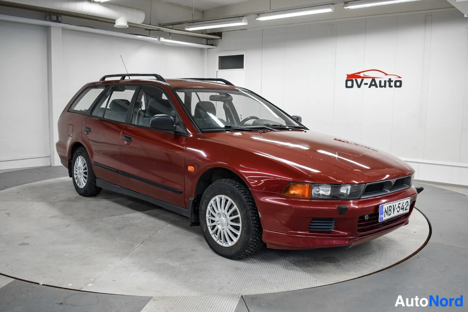 Etuviisto, nokka oikealle: Punainen Mitsubishi Galant -farmari kolmenvartalanäkymästä sisätiloissa, rekisteritunnus.