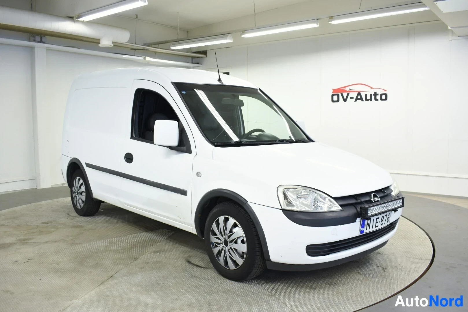 Etuviisto, nokka vasemmalle: Valkoinen pakettiauto, jossa on rekisterikilpi, näkyy etuviistosta oikealta edestäpäin valaistussa näyttelytilassa. Autossa on musta lisävalopa