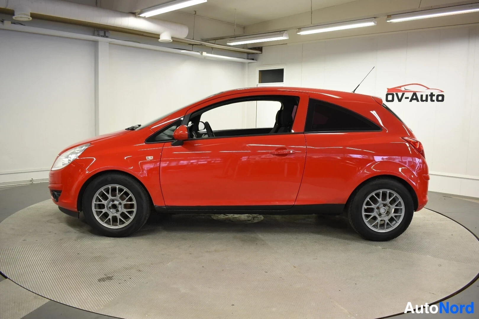 Sivulta: Punainen Opel Corsa kuvattuna vasemmalta sivulta. Autossa on kaksi ovea ja hopeanväriset vanteet.