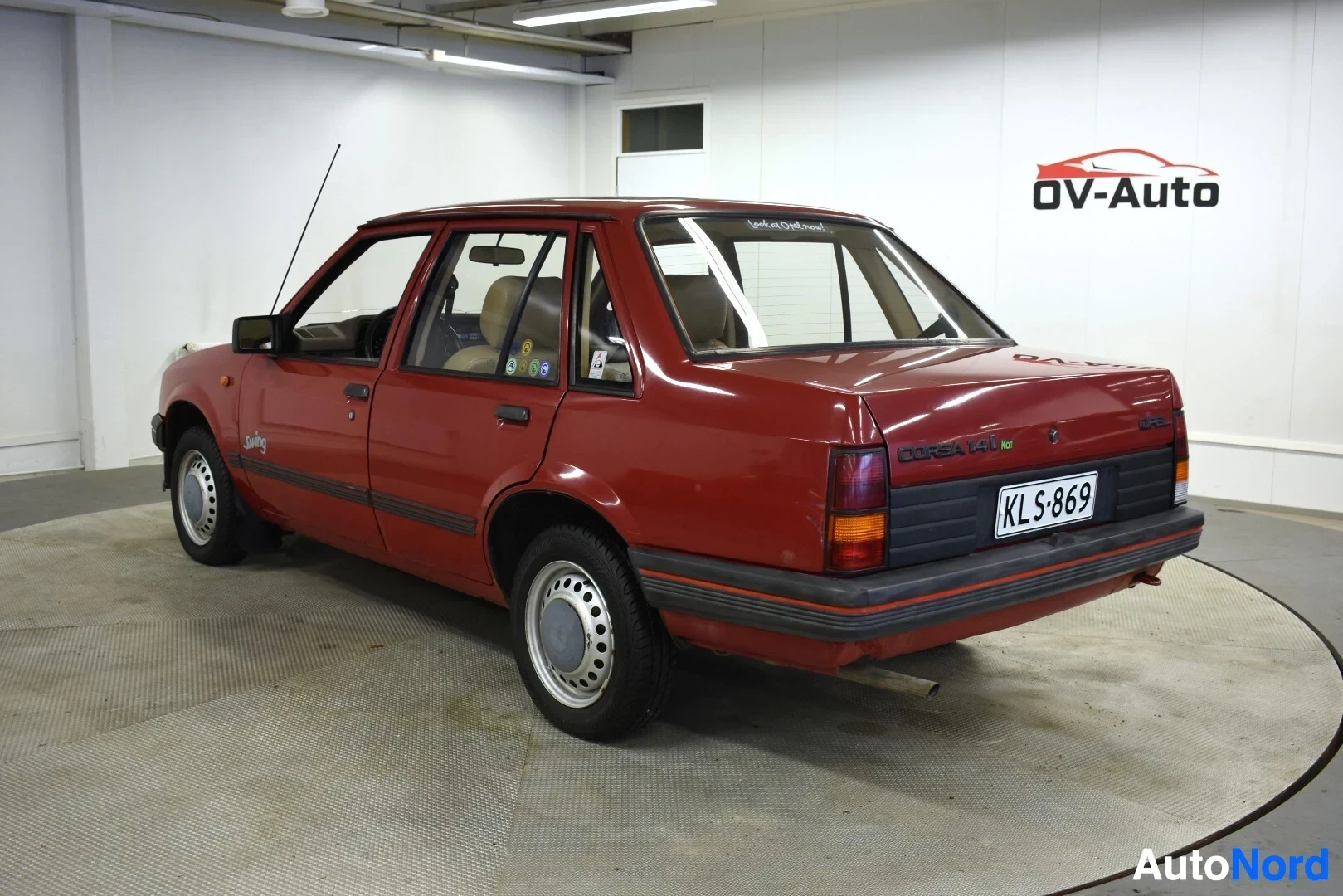 Takaviisto: Punainen henkilöauto kuvattuna takaviistosta oikealta puolelta. Auton rekisterikilvessä lukee "". Takalasissa on tarra, jossa lukee "Licensed to Opel Cor