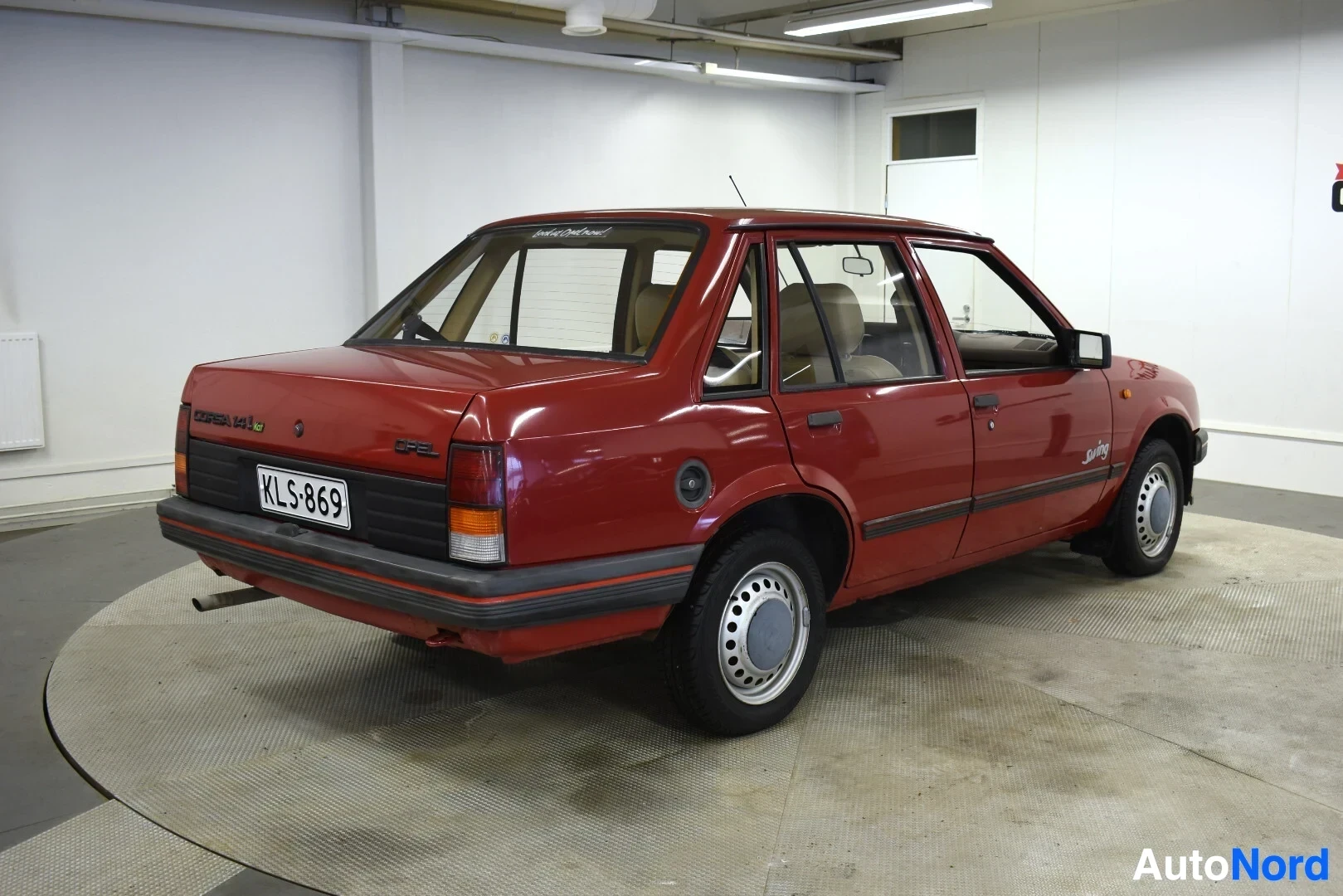 Takaviisto: Punainen henkilöauto kuvattuna takaviistosta vasemmalta puolelta. Auton rekisterikilvessä lukee "". Takalasissa on tarra, jossa lukee "Licensed to Opel C