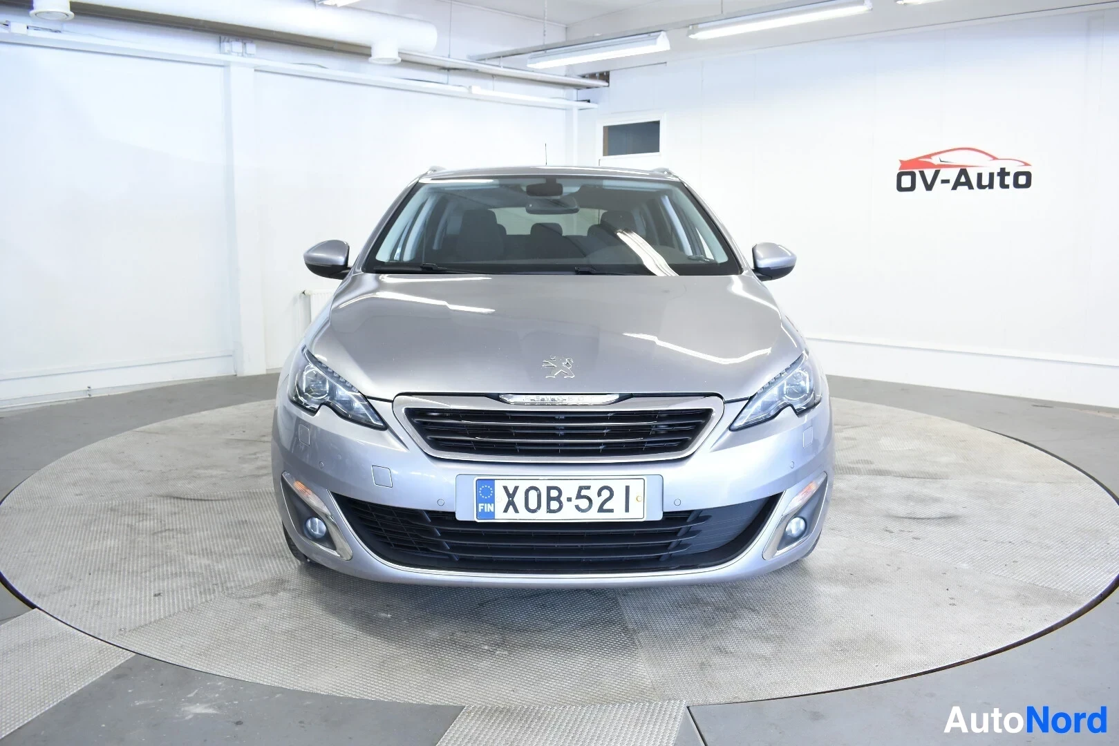 Edestä: Harmaa farmariauto edestä kuvattuna, ajovalot ja maski näkyvissä. Autossa on sumuvalot ja rekisterikilpi.
