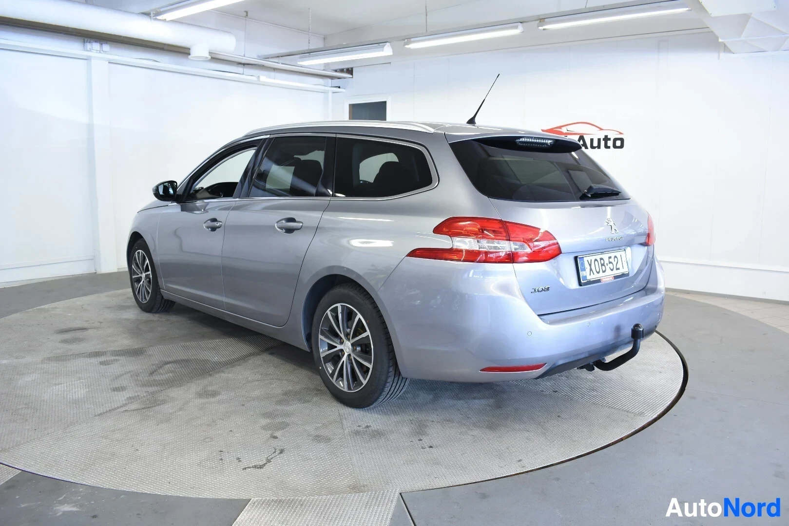 Takaviisto: Hopeanvärinen Peugeot 308 SW takaviistosta oikealta kuvattuna, kuva otettu hieman matalalta. Ajoneuvossa on vetokoukku ja kattokiskot.