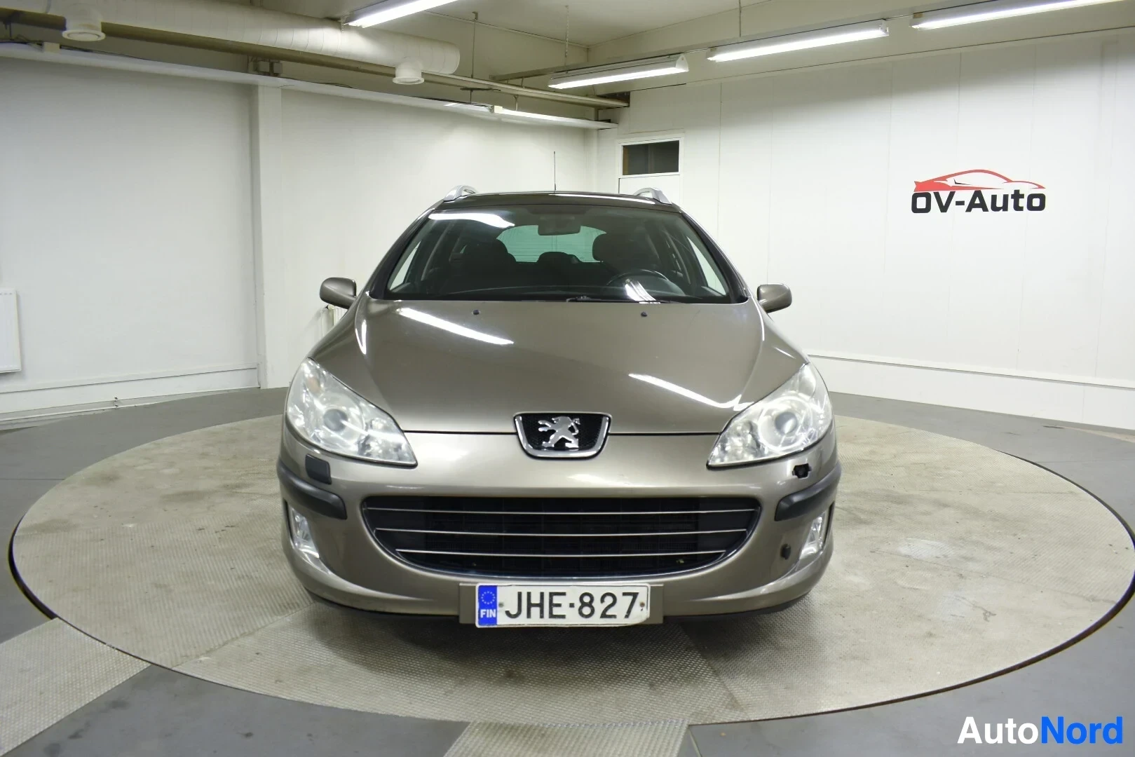 Edestä: Beigen värinen Peugeot 407 farmariauto kuvattu suoraan edestä sisätilassa. Auton rekisterikilvessä lukee ""