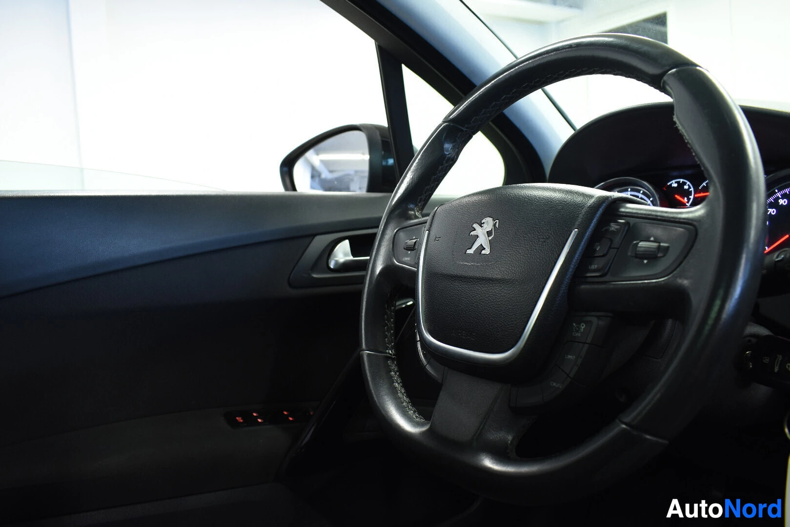 Ohjaamo: Sisäkuva, jossa näkyy auton ohjauspyörä ja osa kuljettajan ovesta. Ohjauspyörässä on Peugeot-logo.