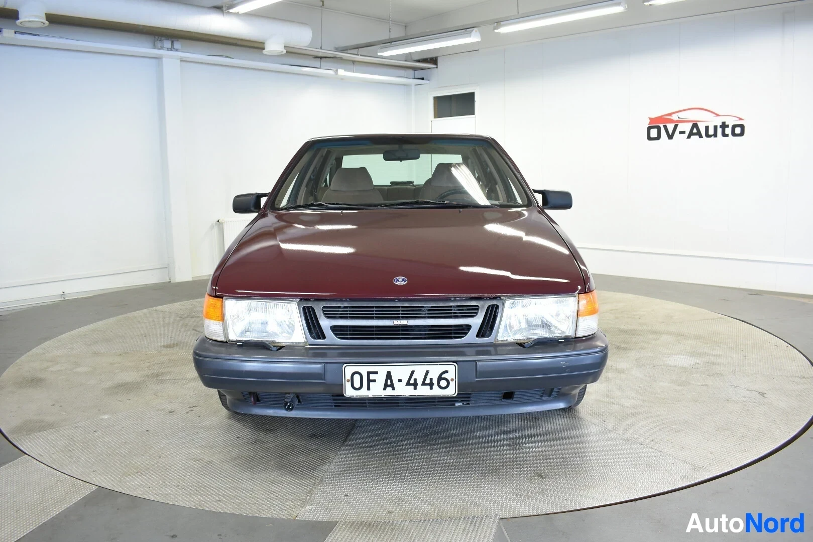 Edestä: Burgundinpunainen henkilöauto edestä kuvattuna, rekisterikilpi. Autossa on suorakulmaiset etuvalot ja säleikkö, jonka yläosassa on Saab-logo. Auto on py