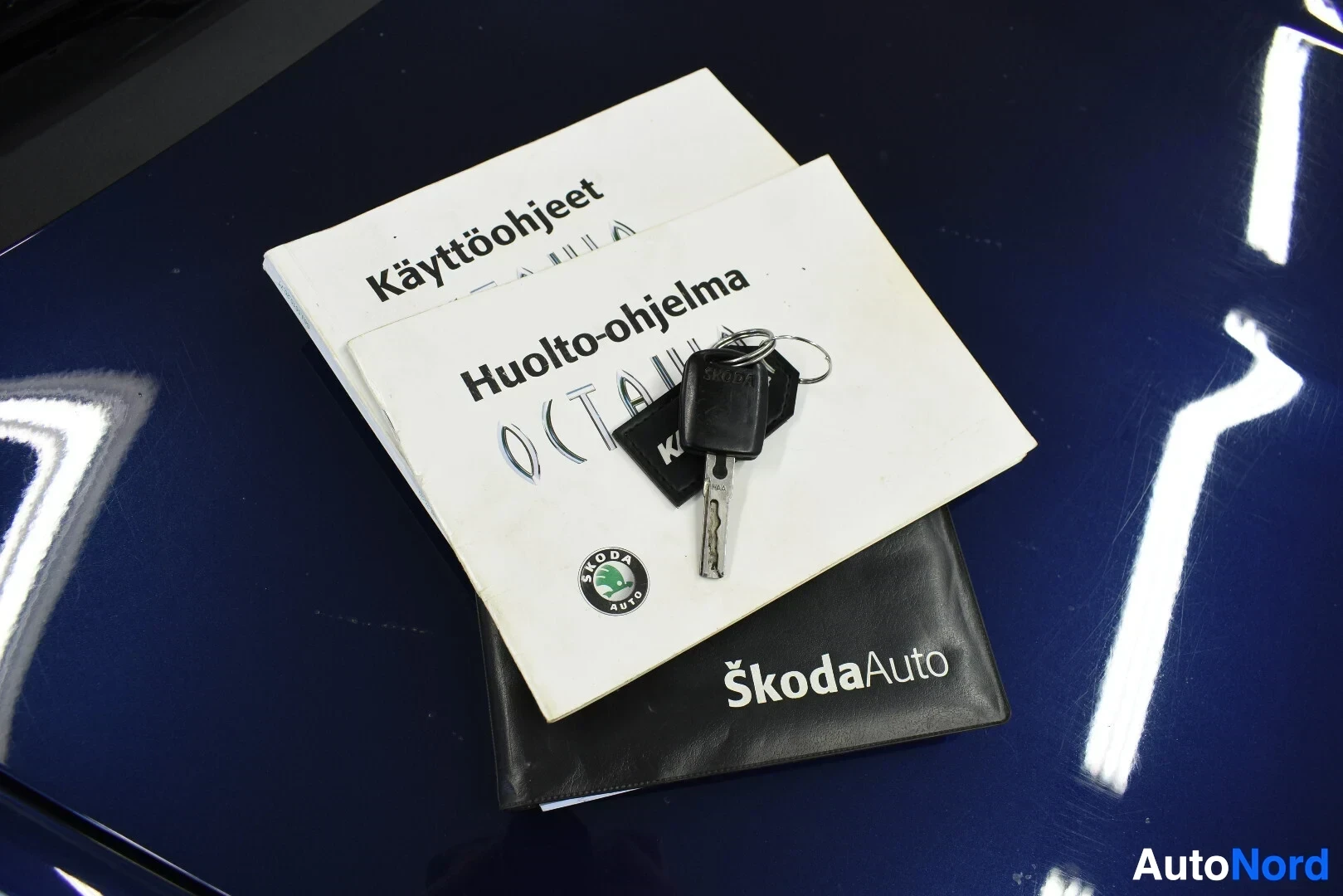 Yksityiskohta: Käyttöohjeet- ja Huolto-ohjelma-nimiset kirjat ja Skoda-avain nipussa mustan kotelon päällä sinisellä pinnalla.