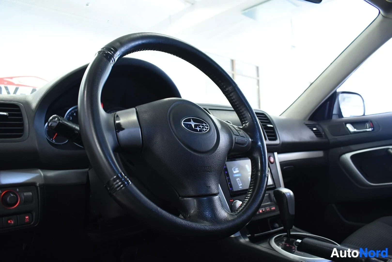 Ohjaamo: Kuvassa näkyy auton ohjauspyörä, jossa on Subaru-logo ja monitoiminäppäimet. Kojelauta ja osittain vaihdekeppi ovat myös kuvassa.