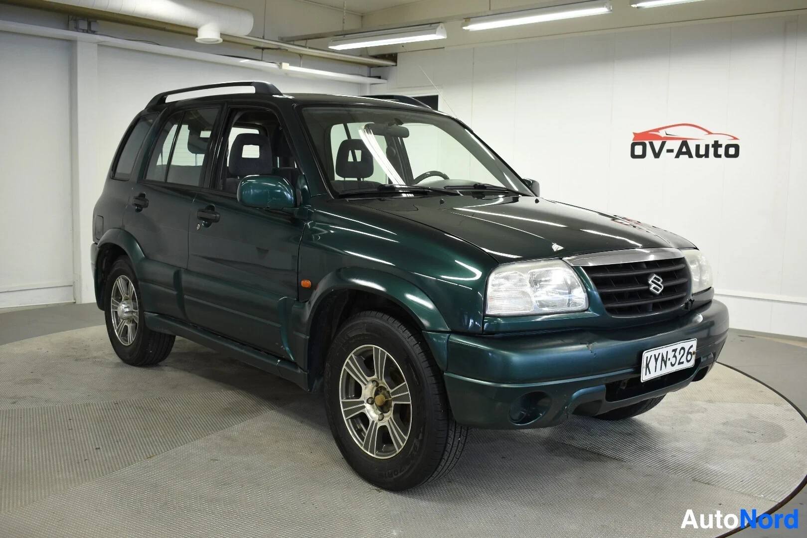 Etuviisto, nokka vasemmalle: Tummanvihreä Suzuki Grand Vitara on pysäköity halliin, sen etuosa ja oikea kylki näkyvät kuvassa. Autossa on kattokaiteet ja kevytmetallivanteet. Rekis