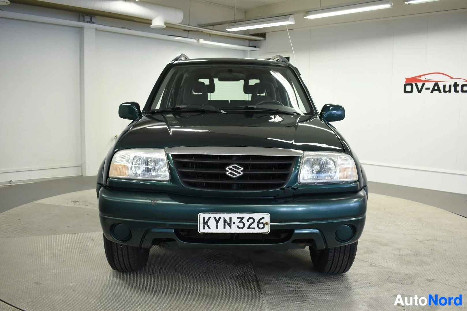 Edestä: Tummanvihreä Suzuki Grand Vitara on kuvattu edestäpäin hallissa. Autossa on kattokaiteet. Rekisterikilpi "" on kiinnitetty etupuskuriin