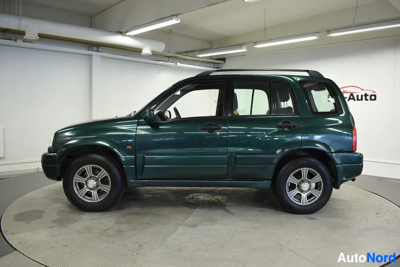 Sivulta: Tummanvihreä Suzuki Grand Vitara on kuvattu vasemmalta kyljeltä hallissa. Autossa on kattokaiteet ja kevytmetallivanteet.
