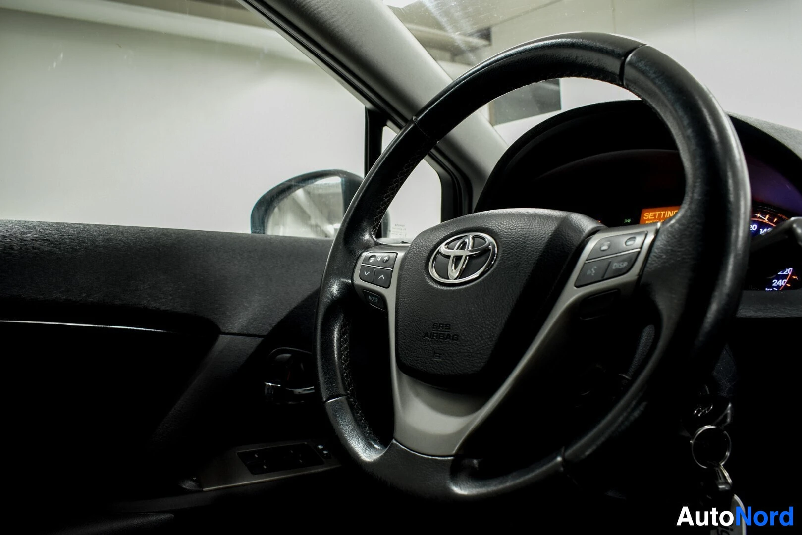 Ohjaamo: Lähikuva ohjauspyörästä ja osasta kuljettajan oven sisäverhoilua. Ohjauspyörän keskellä on Toyota-logo.