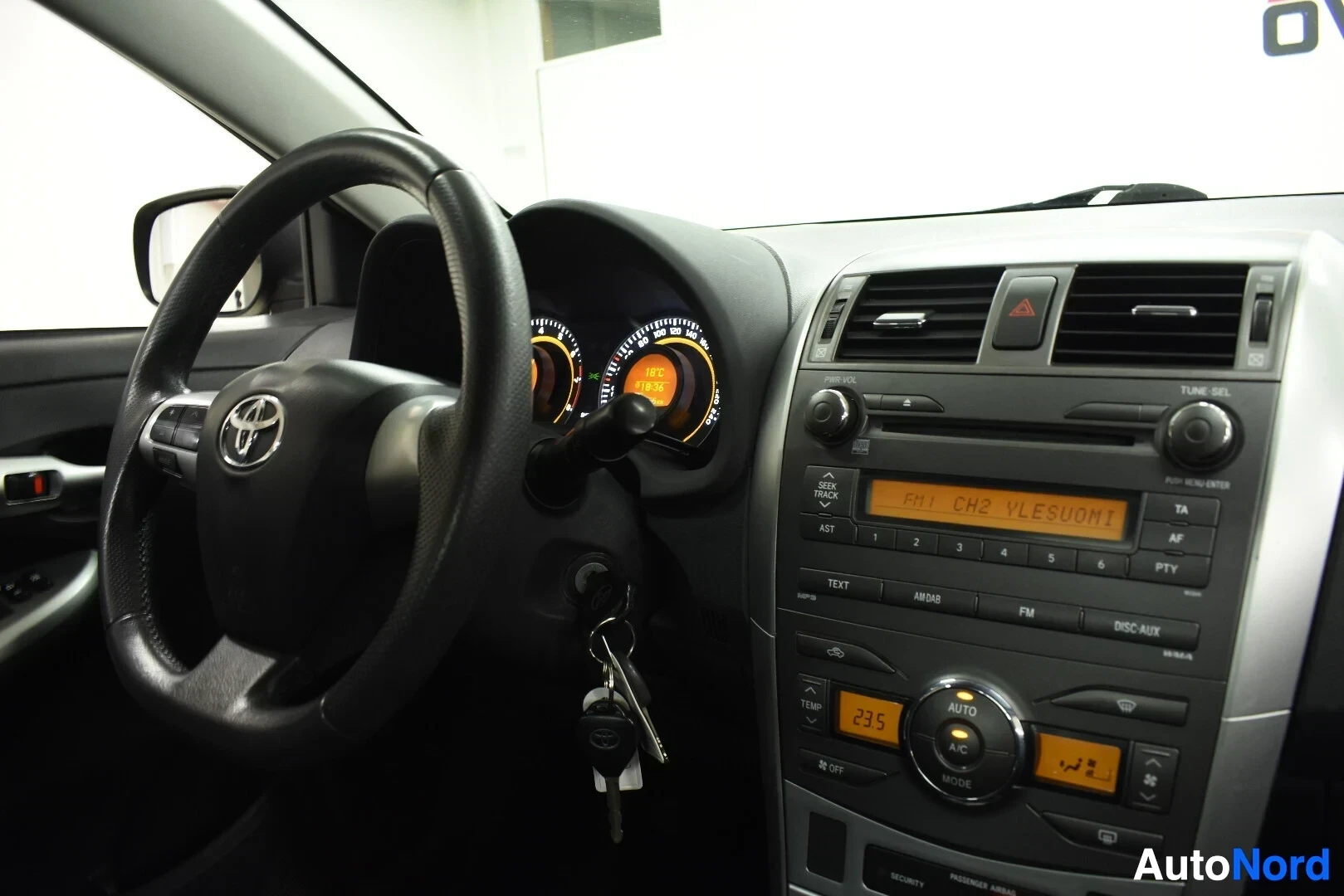 Ohjaamo: Kojelauta ja ohjauspyörä, jossa on Toyota-logo. Keskikonsolissa on radio, jossa lukee "FM1 CH2 YLESUOMI", ja automaattinen ilmastointipaneeli, jossa näkyy lämpö