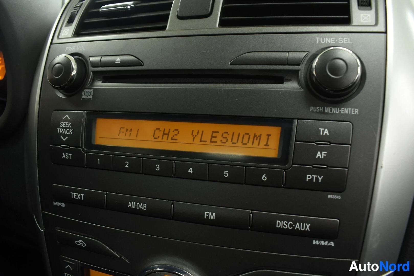 Yksityiskohta: Lähikuva auton radiosta, jossa näytöllä lukee "FM1 CH2 YLESUOMI". Radion painikkeet ovat näkyvissä, mukaan lukien "TUNE-SEL", "PUSH MENU-ENTER", "AST", "SEEK TR