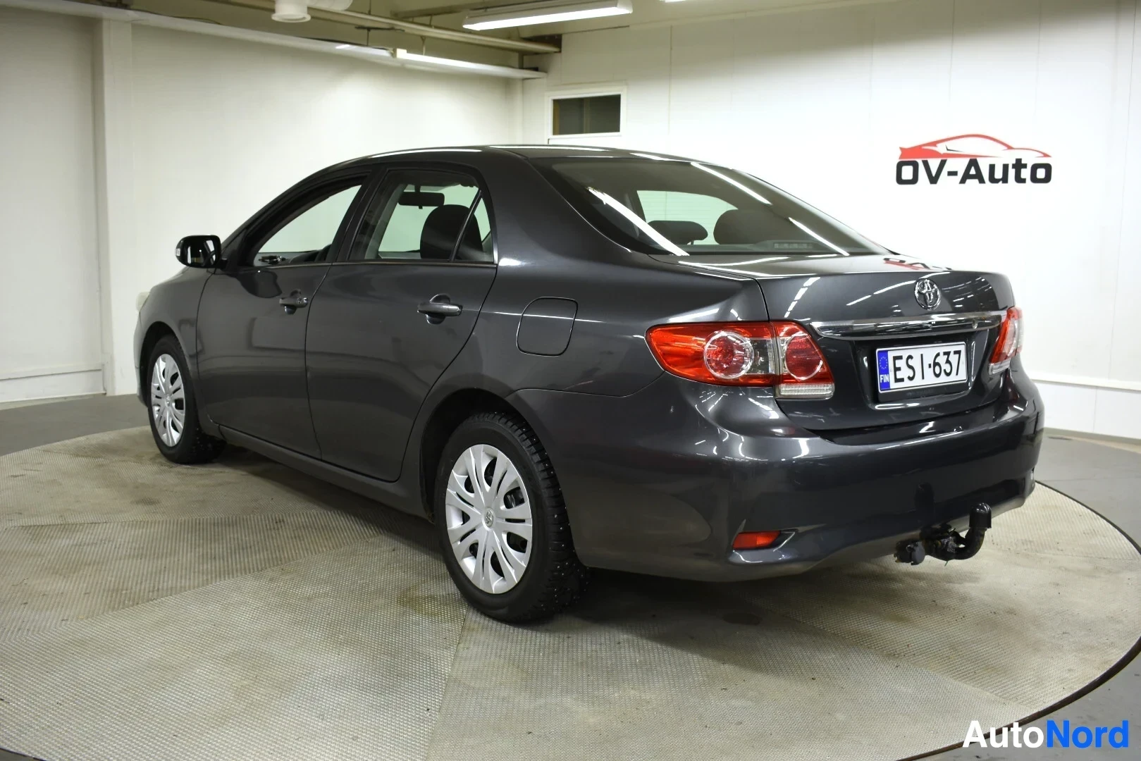 Takaviisto: Tummanharmaa sedan-tyyppinen auto kuvattuna takavasemmalta kolmeneljänneskulmasta sisätiloissa.