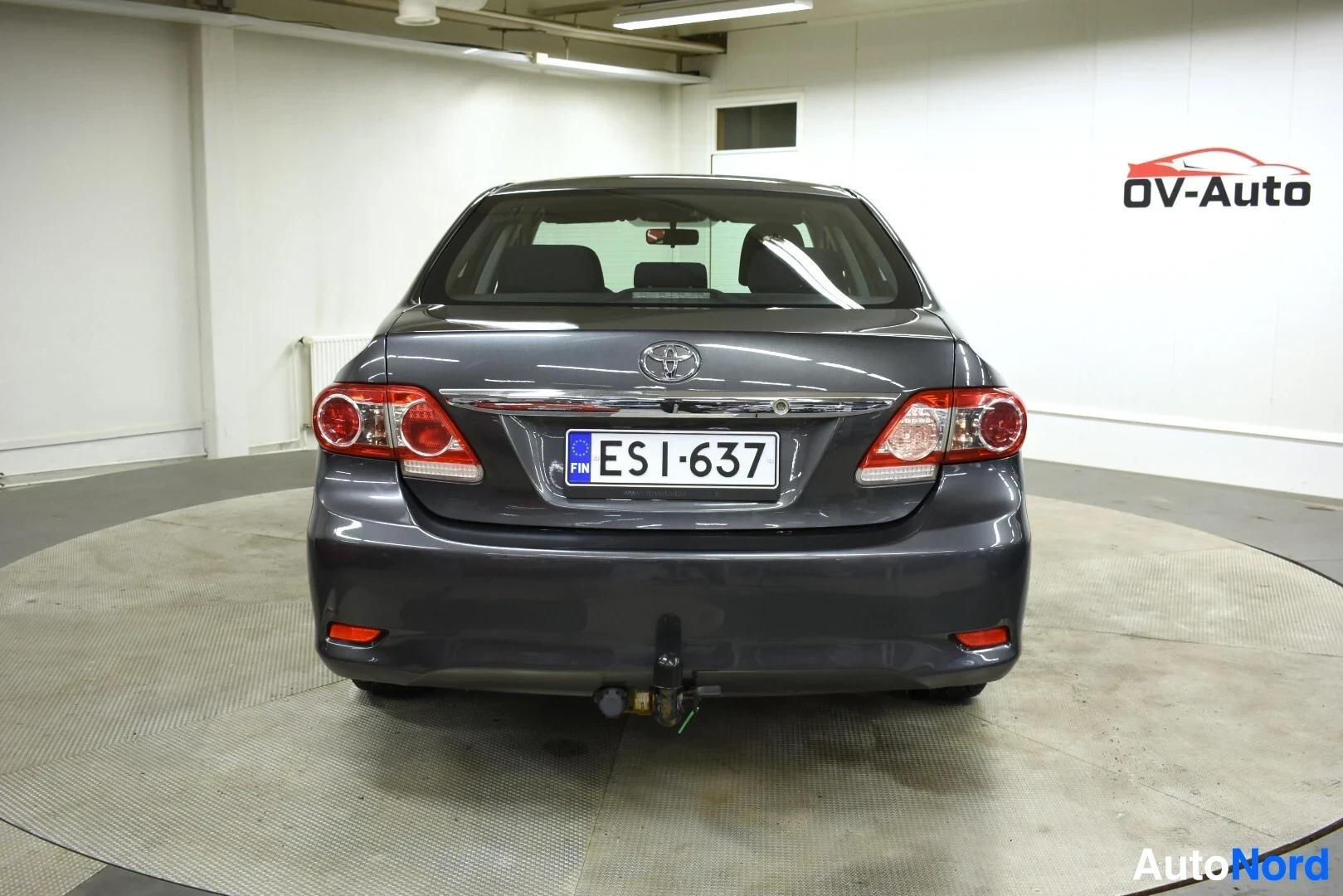Takaa: Tummanharmaa sedan-tyyppinen auto kuvattuna suoraan takaa sisätiloissa.