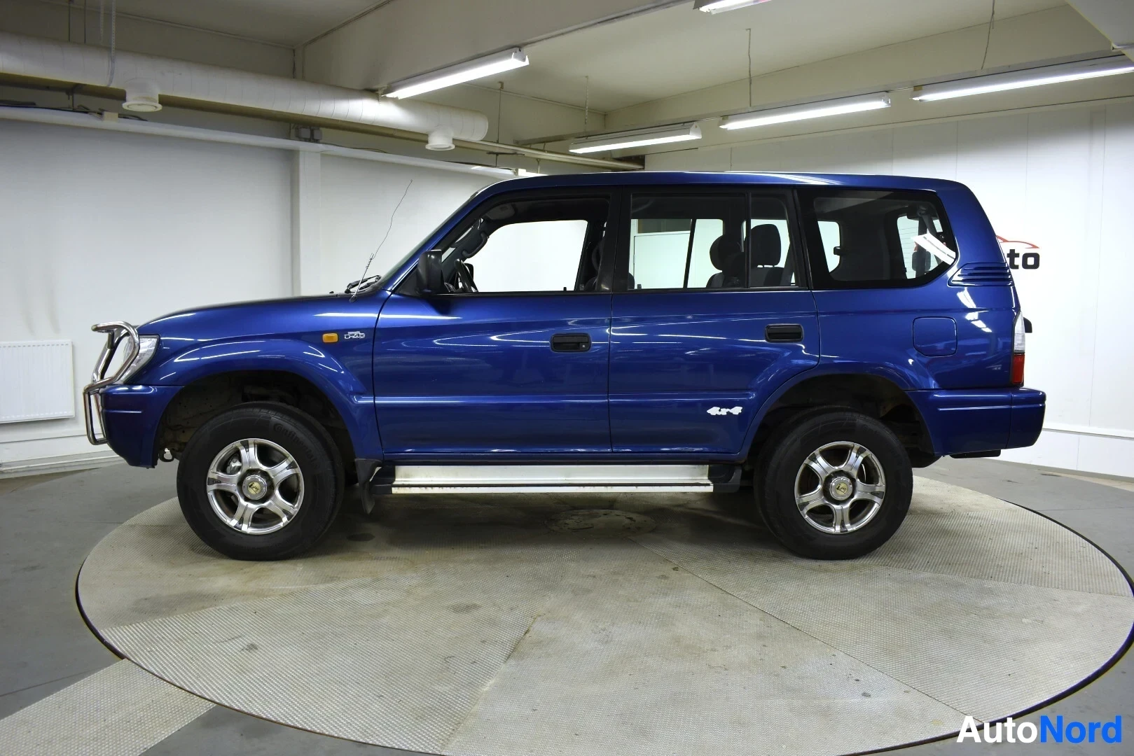 Sivulta: Sinisestä maastoautosta otettu kuva vasemmalta puolelta. Autossa on astinlaudat ja takapyörän lähellä lukee "4x4".