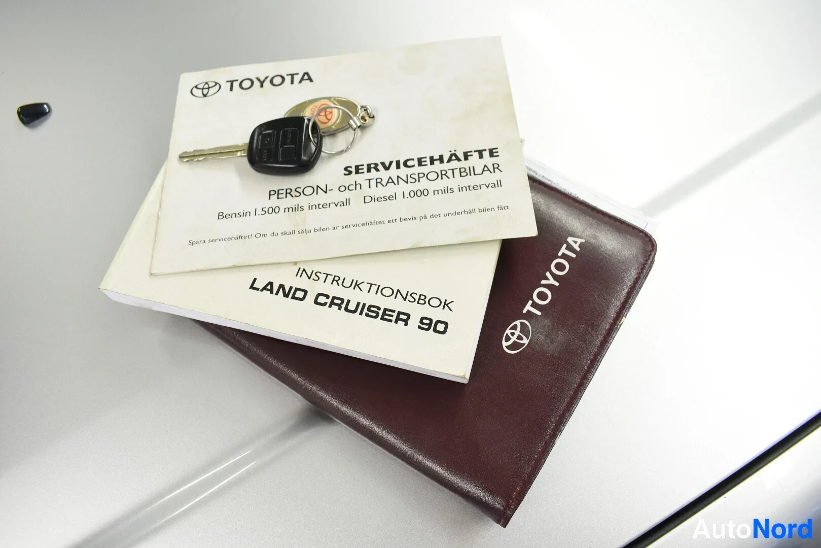Yksityiskohta: Kuva auton hopeisella konepellillä makaavista dokumenteista ja avaimesta. Päällimmäisenä on Toyota-merkkinen avain, jonka alla on huoltokirja "SERVICEHÄFTE" ja