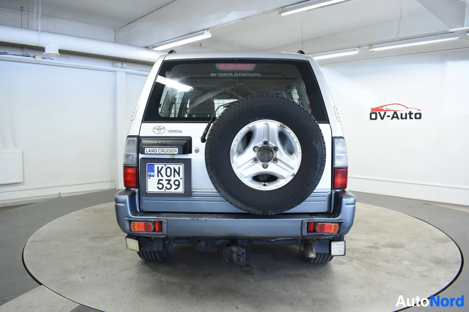 Takaa: Harmaa maastoauto takaa kuvattuna, ajoneuvossa on vetokoukku, ulkoinen vararengas takaluukussa ja kattokaiteet. Rekisterikilpi "KON 539" on luettavissa.