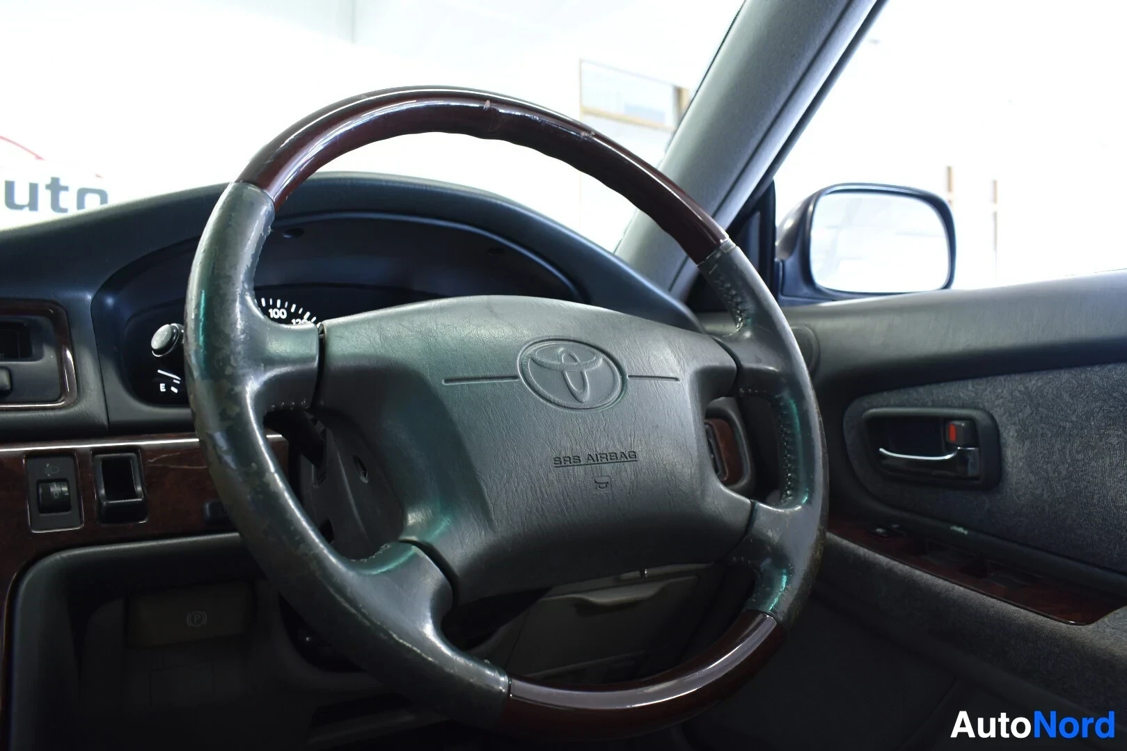 Yksityiskohta: Lähikuva auton ohjauspyörästä, jossa on puunvärisiä yksityiskohtia ja Toyota-logo keskellä. Matkustajan puoleinen sivuovi on näkyvissä oikealla.