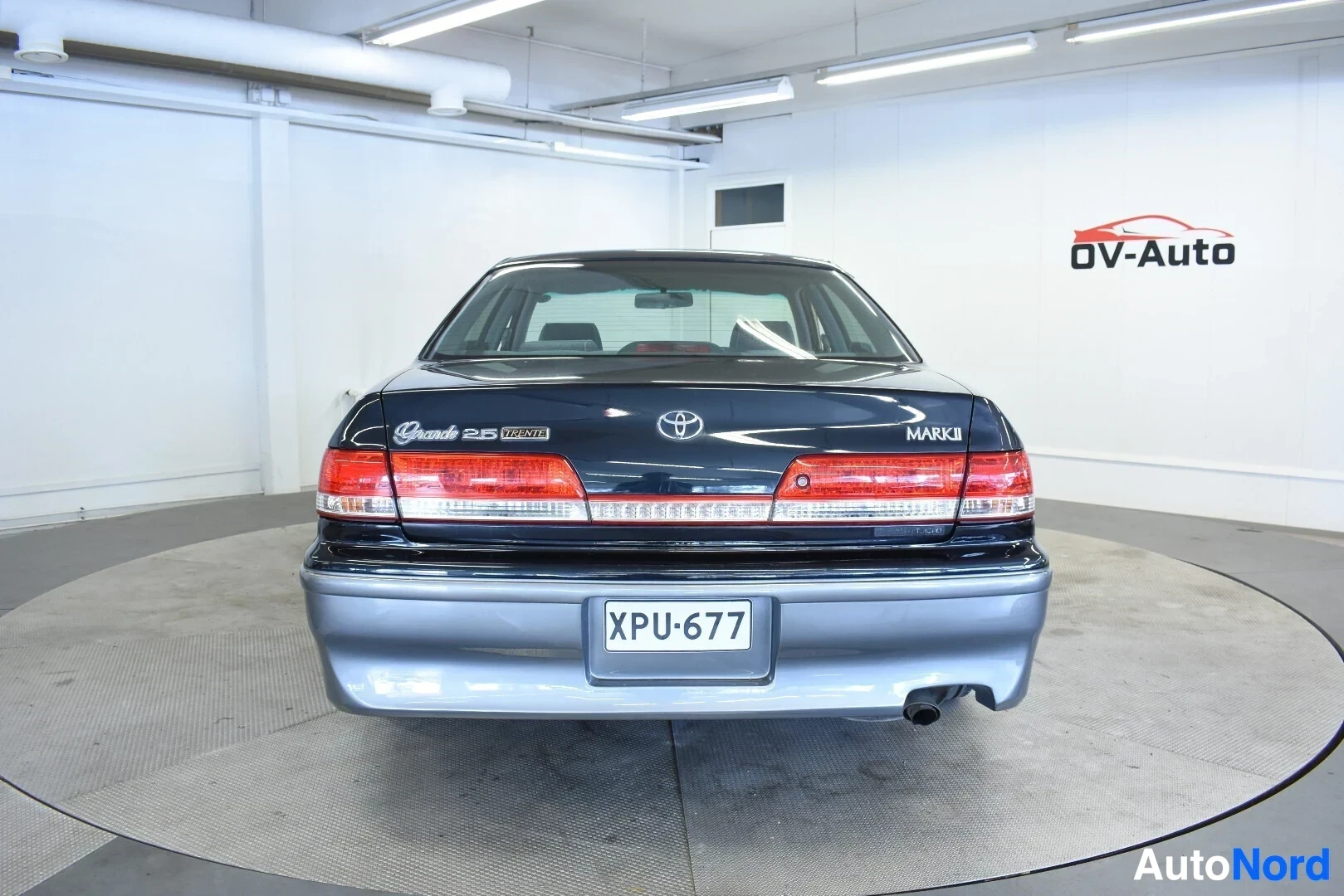 Takaa: Tummanharmaa sedan-tyyppinen auto kuvattuna takaa. Takarekisterikilvessä lukee. Takaluukussa on merkinnät "Grande 2.5 iR-V", "TOYOTA" ja "MARK II"