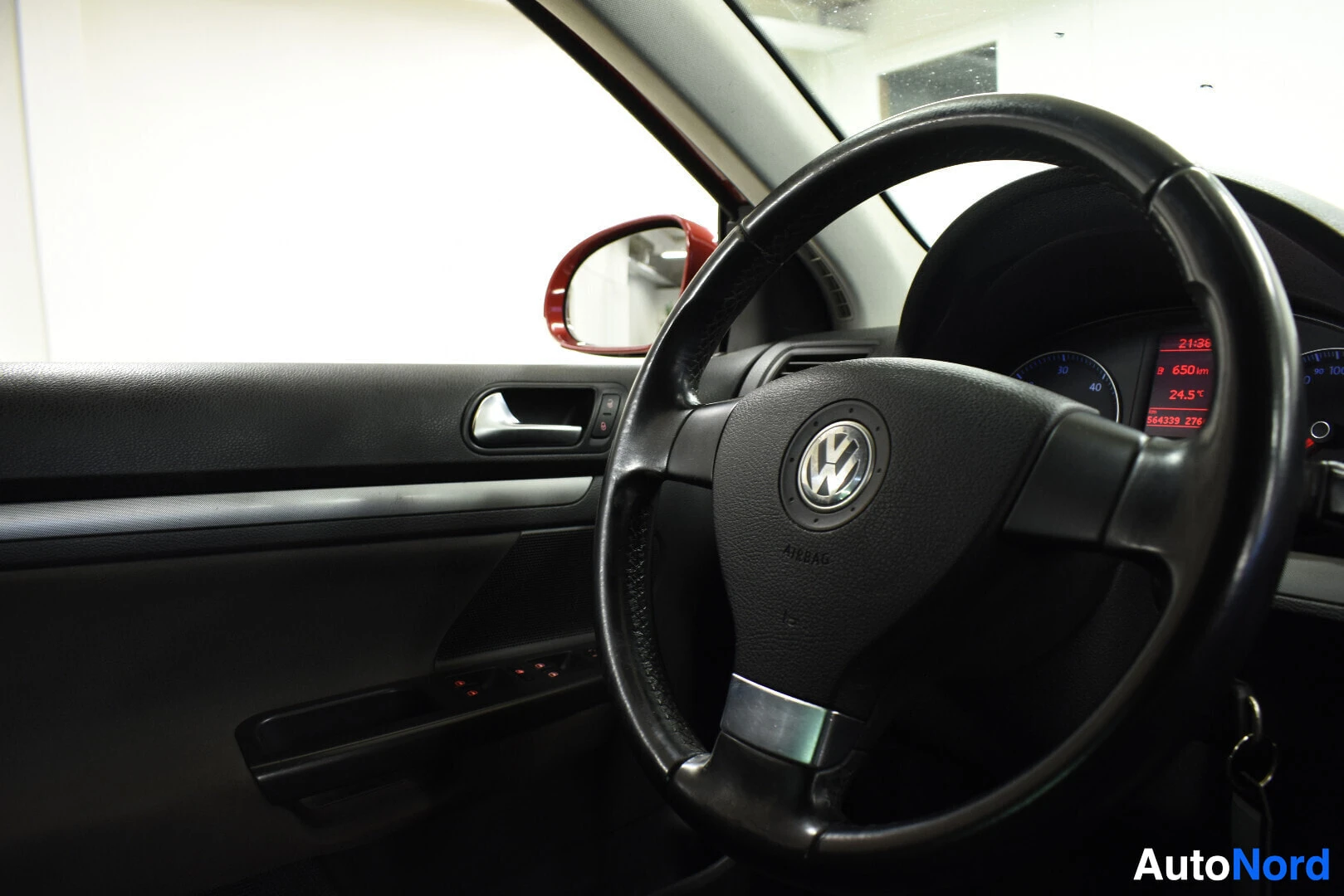Ohjaamo: Lähikuva auton sisätiloista, keskittyen kuljettajan oven ja ohjauspyörän alueelle. Musta ohjauspyörä, jossa on Volkswagenin logo ja teksti "AIRBAG", hallitsee k