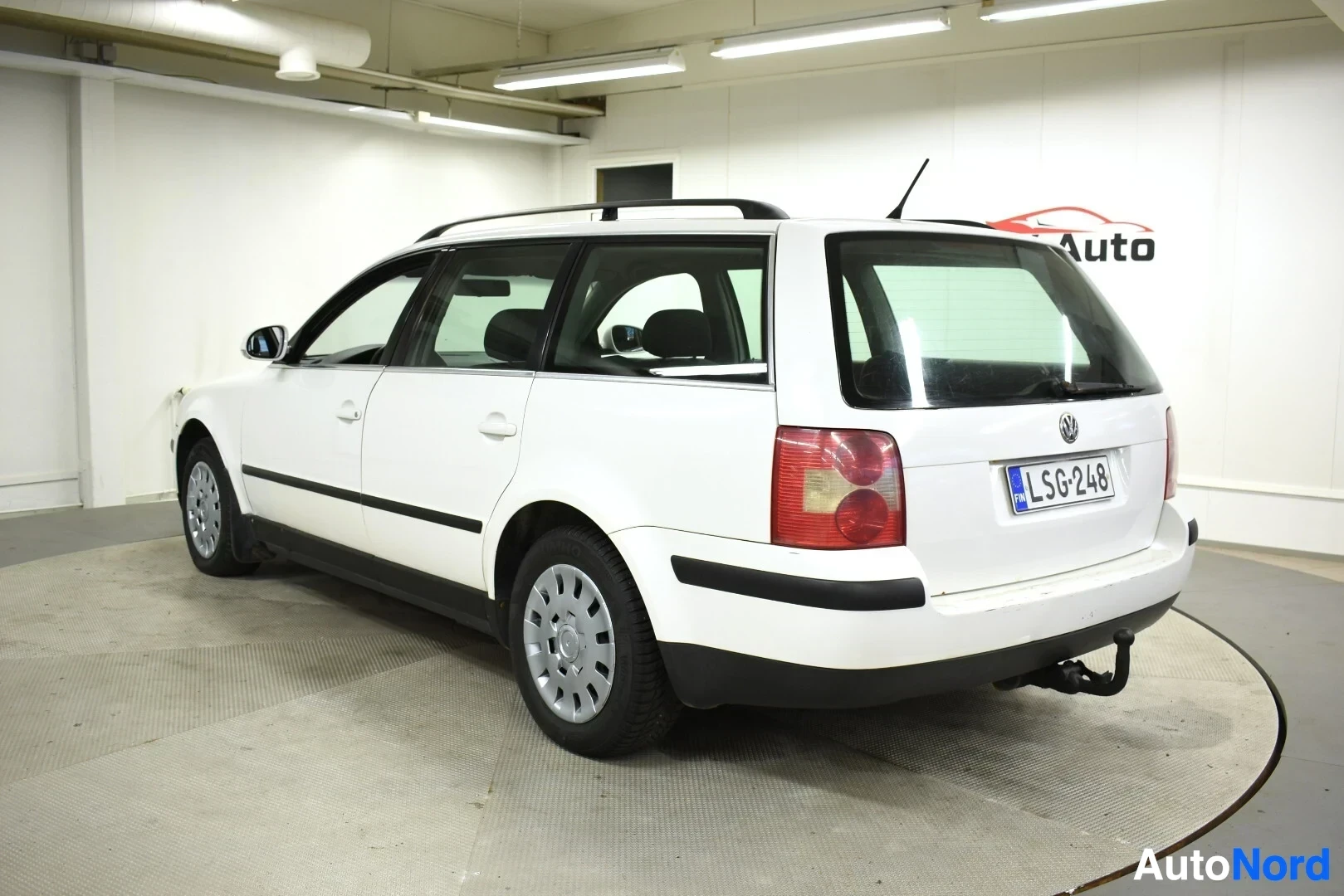 Takaviisto: Valkoinen farmariauto, jossa on mustat kattokaiteet ja vetokoukku, takaoikealta kuvattuna.