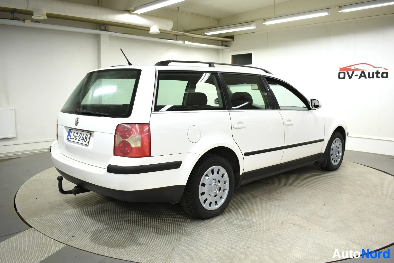 Takaviisto: Valkoinen farmariauto, jossa on mustat kattokaiteet ja vetokoukku, takavasemmalta kuvattuna.