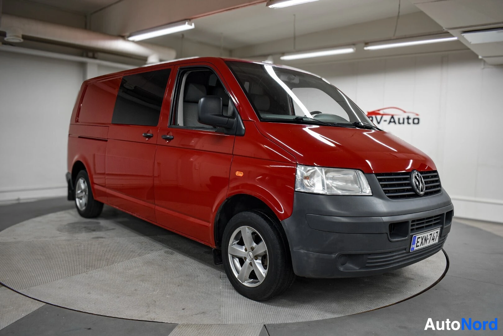 Etuviisto, nokka oikealle: Punainen Volkswagen Transporter -pakettiauto kuvattuna kolmannenvälinen etukulmasta vasemmalta, koko ajoneuvo näkyy.