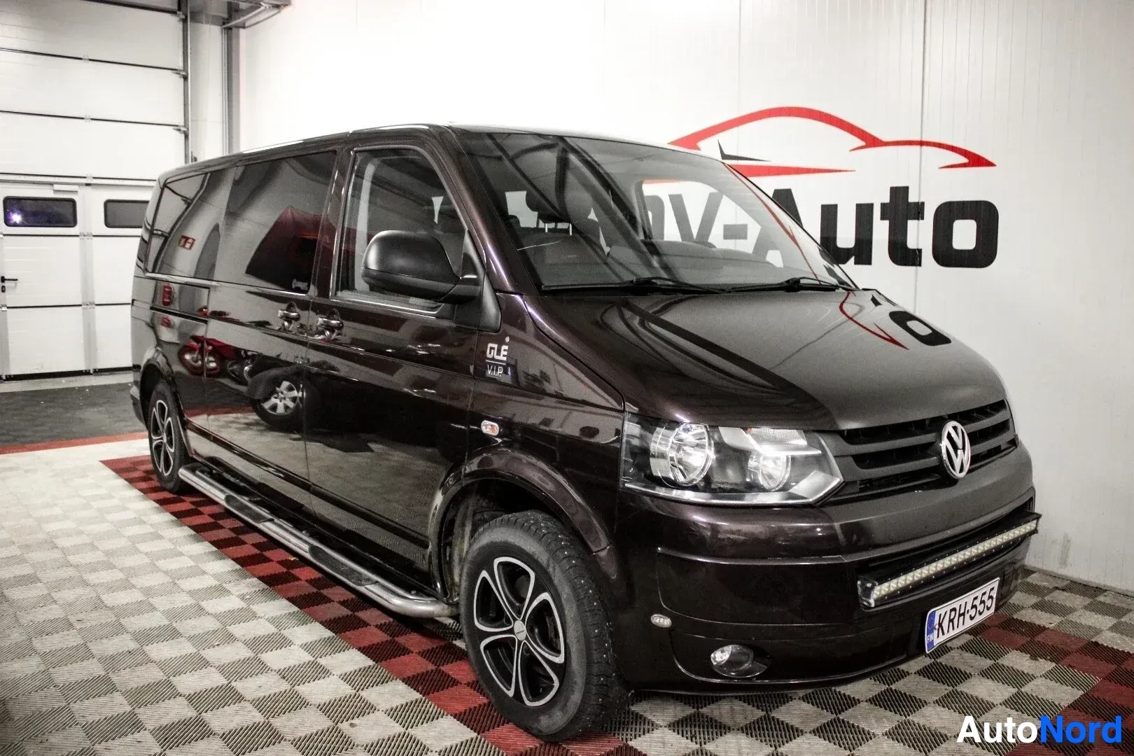 Etuviisto, nokka vasemmalle: Tummanruskea Volkswagen Transporter pakettiauto, jossa on astinlaudat ja lisävalopaneeli, kuvattuna etuviistosta oikealta puolelta autohallissa. Autoss