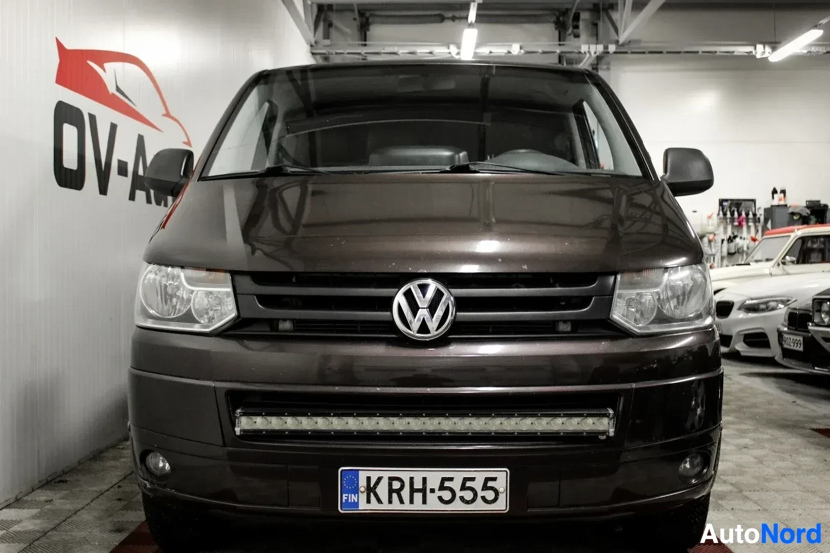 Edestä: Tummanruskea Volkswagen Transporter pakettiauto edestä päin kuvattuna autohallissa. Autossa on keulassa lisävalopaneeli ja rekisterikilpi