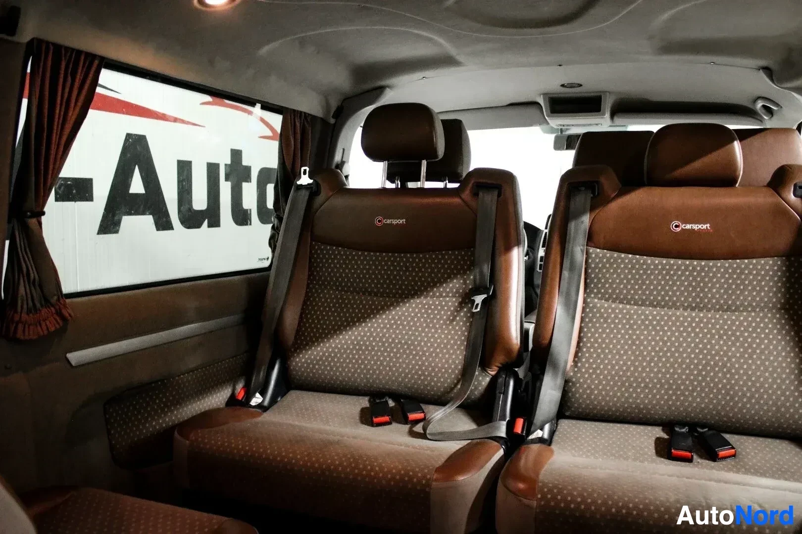 Ohjaamo: Kuva auton takaosasta sisältä, jossa näkyy kaksi ruskea-harmaata istuinta ja oven ikkunasta auton ulkopuolella oleva "Auto"-tekstin osa.