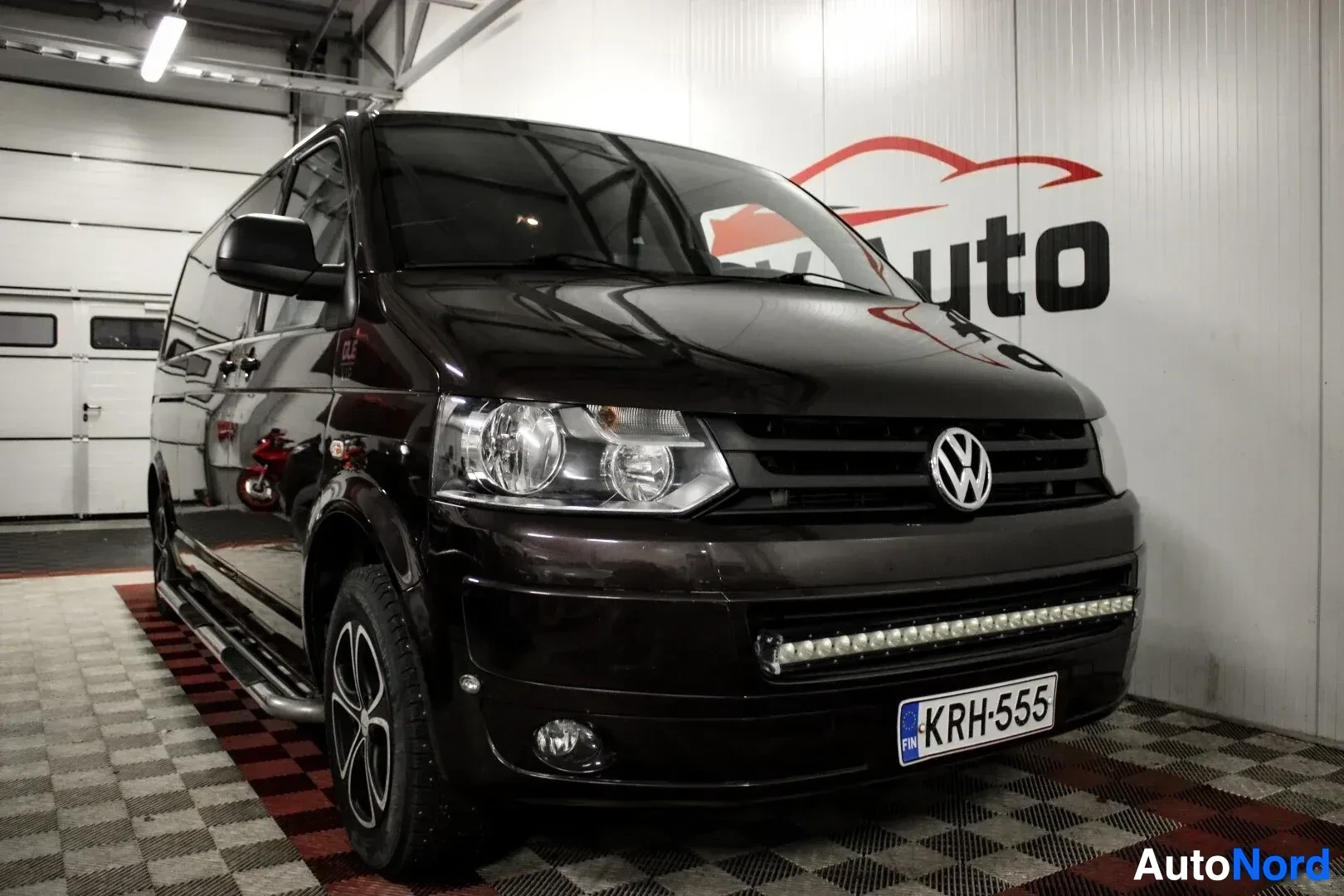 Etuviisto, nokka oikealle: Tummanruskea Volkswagen Transporter pakettiauto, jossa on astinlaudat ja lisävalopaneeli, kuvattuna etuviistosta vasemmalta puolelta autohallissa. Autoss