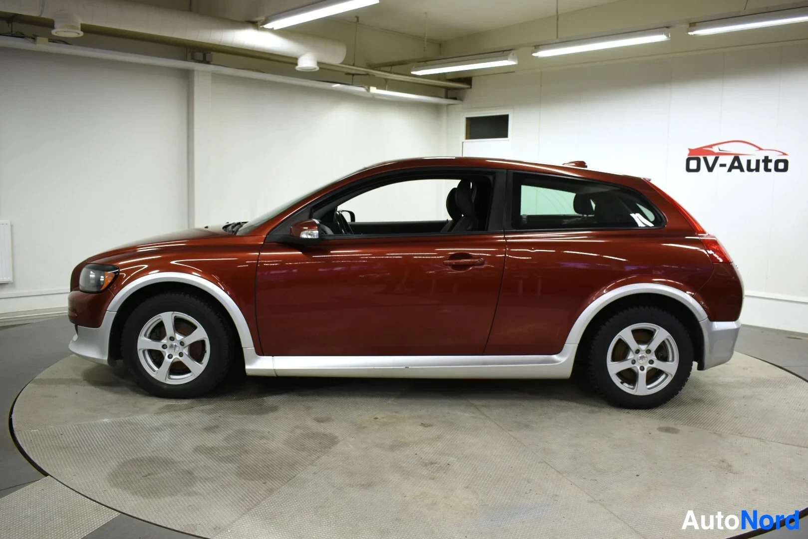 Sivulta: Oranssinruskea Volvo C30 vasemmalta sivulta kuvattuna. Autossa on vaaleanharmaat pyöränkaaret, helmat ja alahelmat sekä kevytmetallivanteet talvirenkailla.