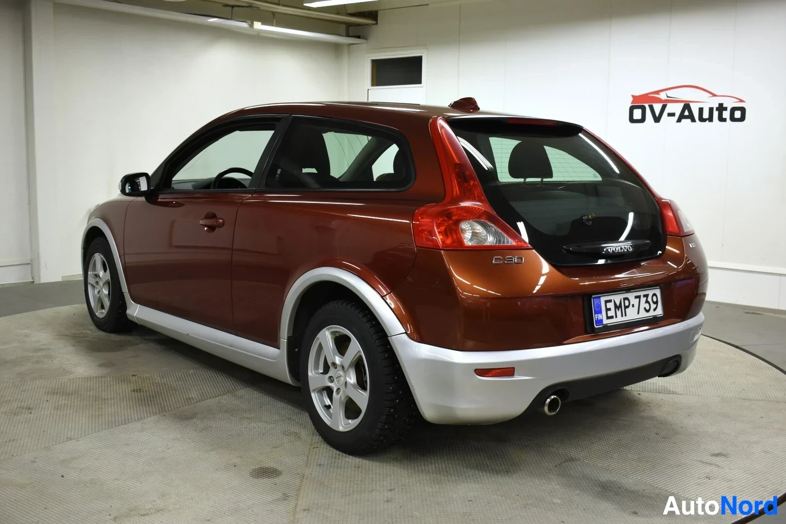 Takaviisto: Volvo C30 -henkilöauto kuvattuna takavasemmalta, näkyvissä takapuskuri, takaluukku ja osa vasemmasta kyljestä. Autossa on ruskea kori ja harmaat helmat. Rekiste