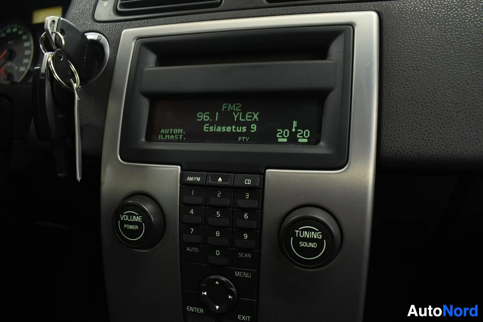 Yksityiskohta: Keskikonsolin lähikuva, jossa näkyy radion ja ilmastoinnin hallintapaneeli. Radion näytöllä lukee "FM2 96.1 YLEX Esiasetus 9 PTV". "VOLUME POWER" ja "TUNING SOU