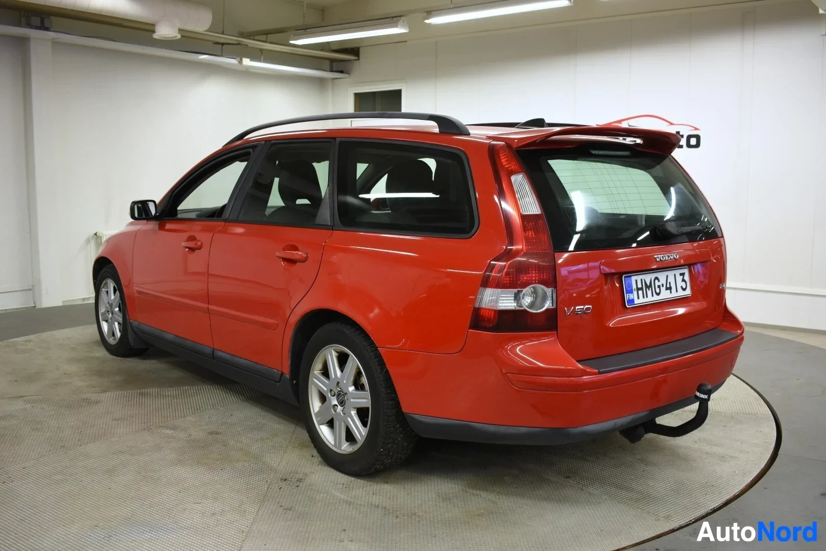 Takaviisto: Oranssinpunainen Volvo V50 farmariauto takavasemmalta kuvattuna. Autossa on kattokiskot ja vetokoukku. Rekisterikilpi on näkyvillä