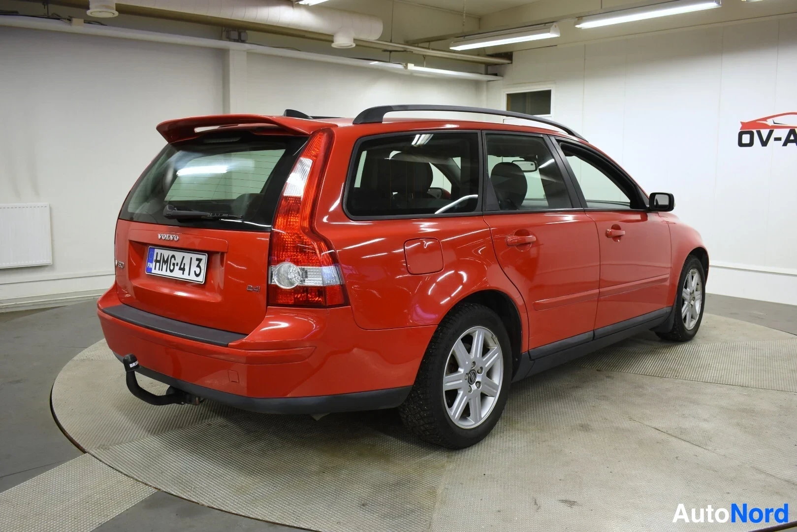 Takaviisto: Oranssinpunainen Volvo V50 farmariauto takaoikealta kuvattuna. Autossa on kattokiskot ja vetokoukku. Rekisterikilpi on näkyvillä