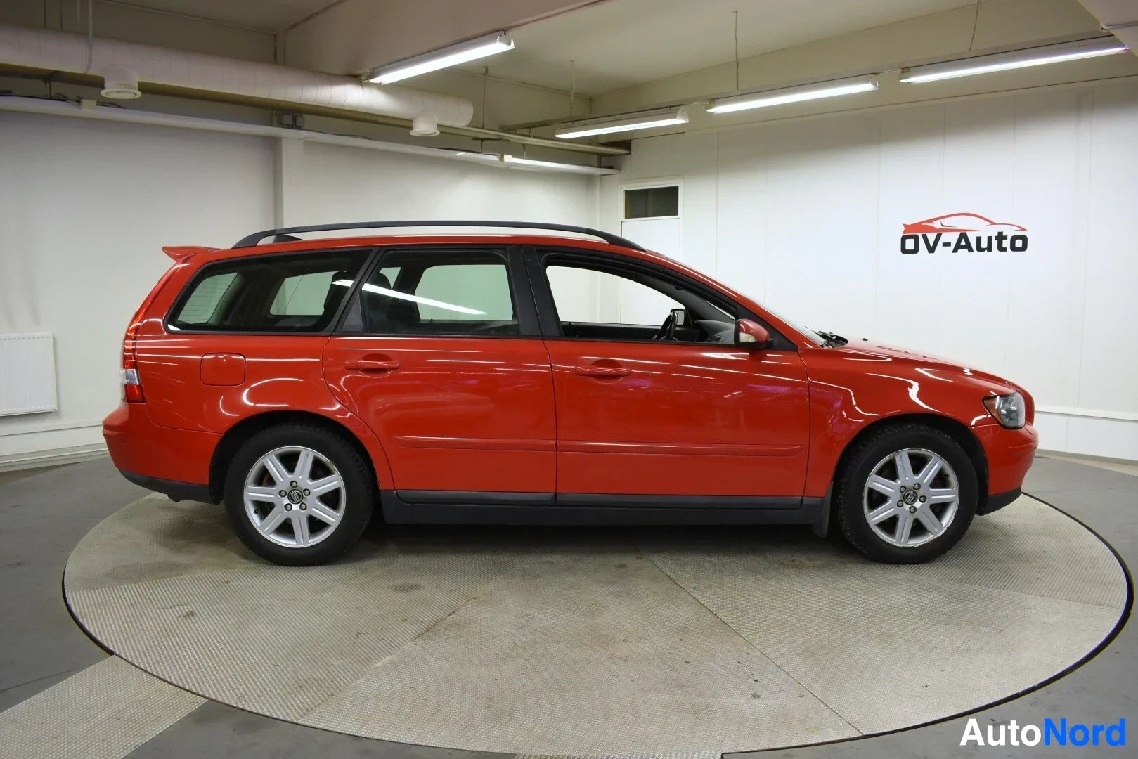 Sivulta: Oranssinpunainen Volvo V50 farmariauto näkyy koko kyljeltään, auton vasen puoli kuvattuna. Autossa on kattokiskot ja kevytmetallivanteet.