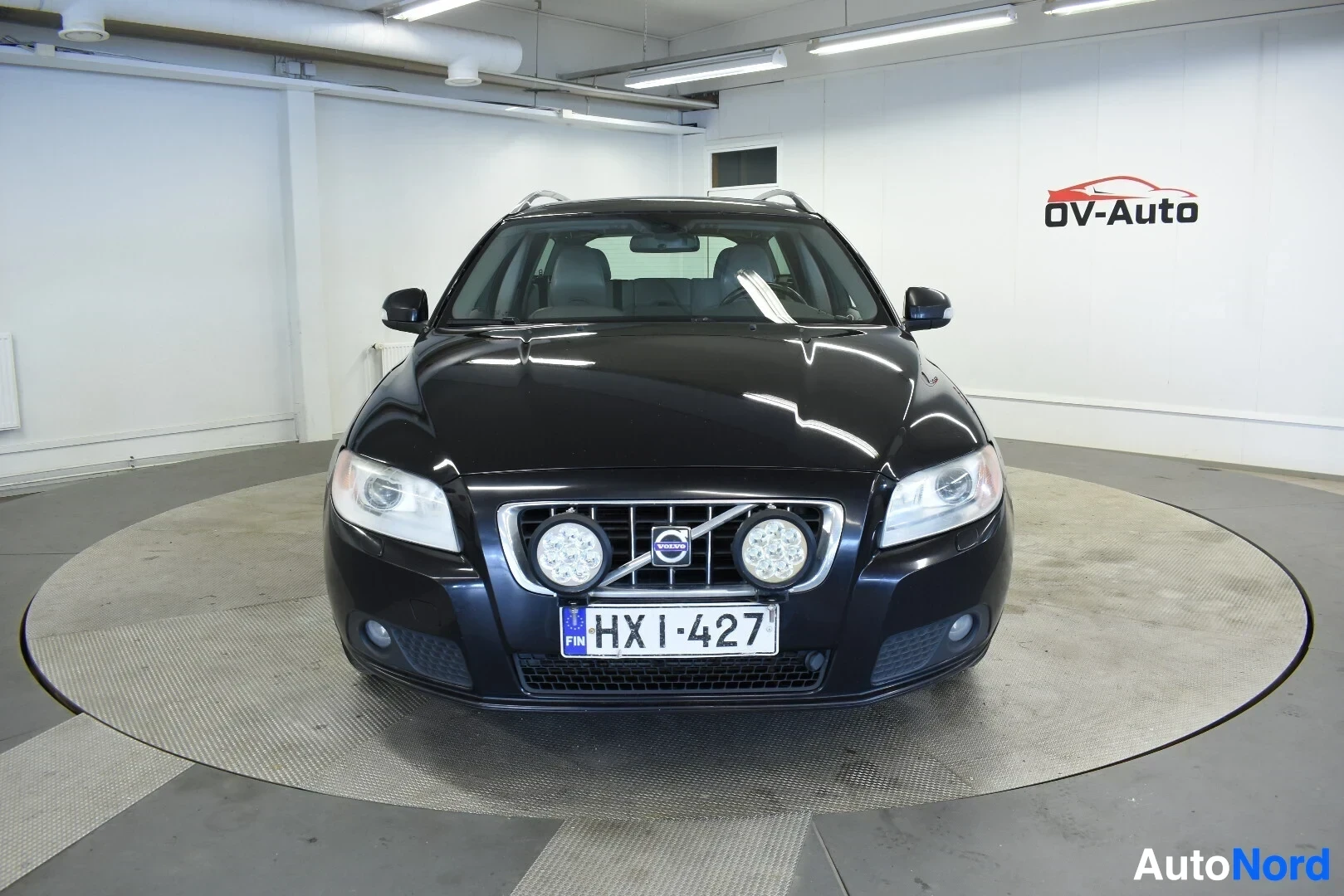 Edestä: Musta Volvo V70 -farmariauto edestä kuvattuna. Autossa on kaksi pyöreää lisävaloa ja rekisterikilpi