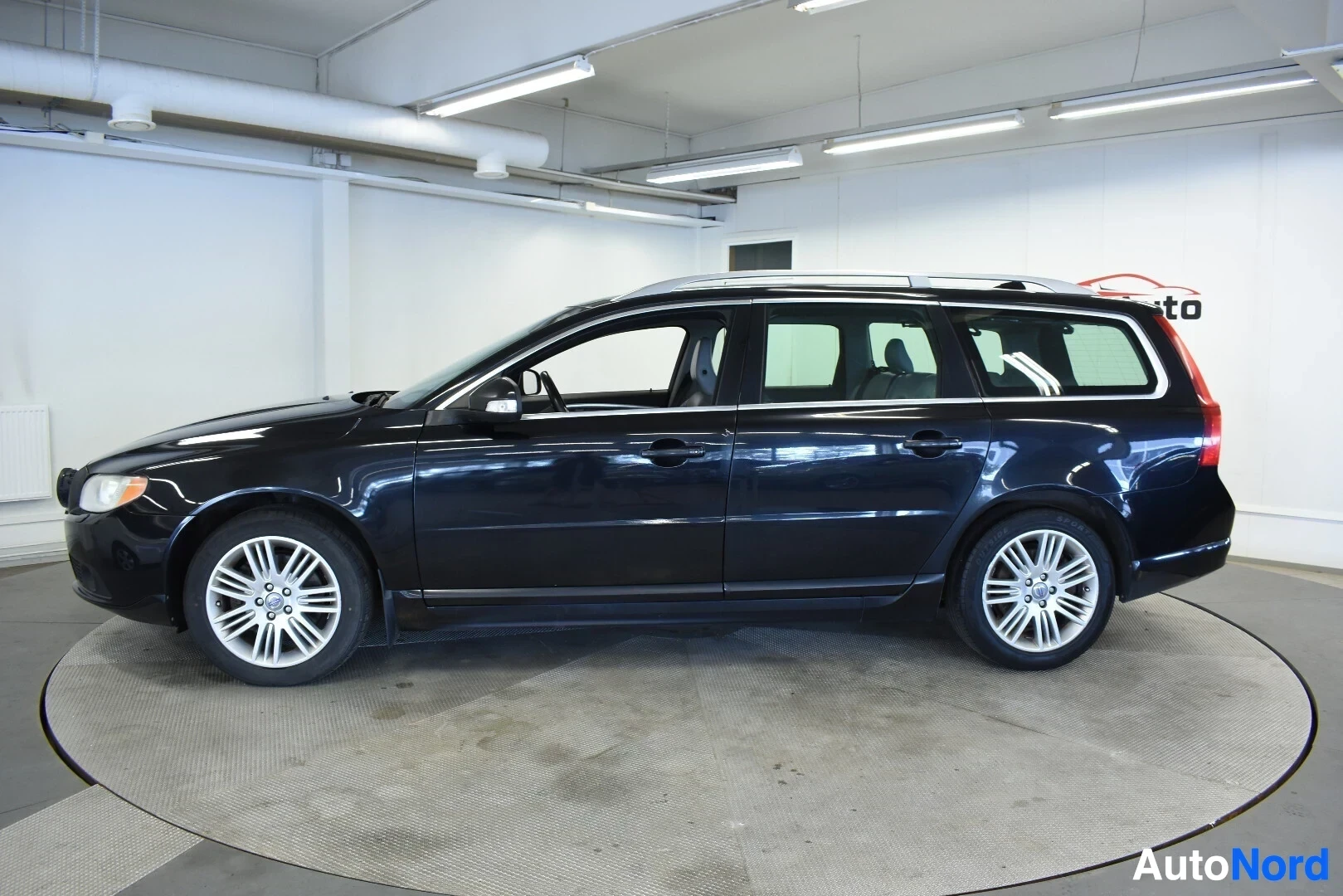 Sivulta: Musta Volvo V70 -farmariauto oikealta sivulta kuvattuna. Autossa on kattokaiteet ja kevytmetallivanteet.