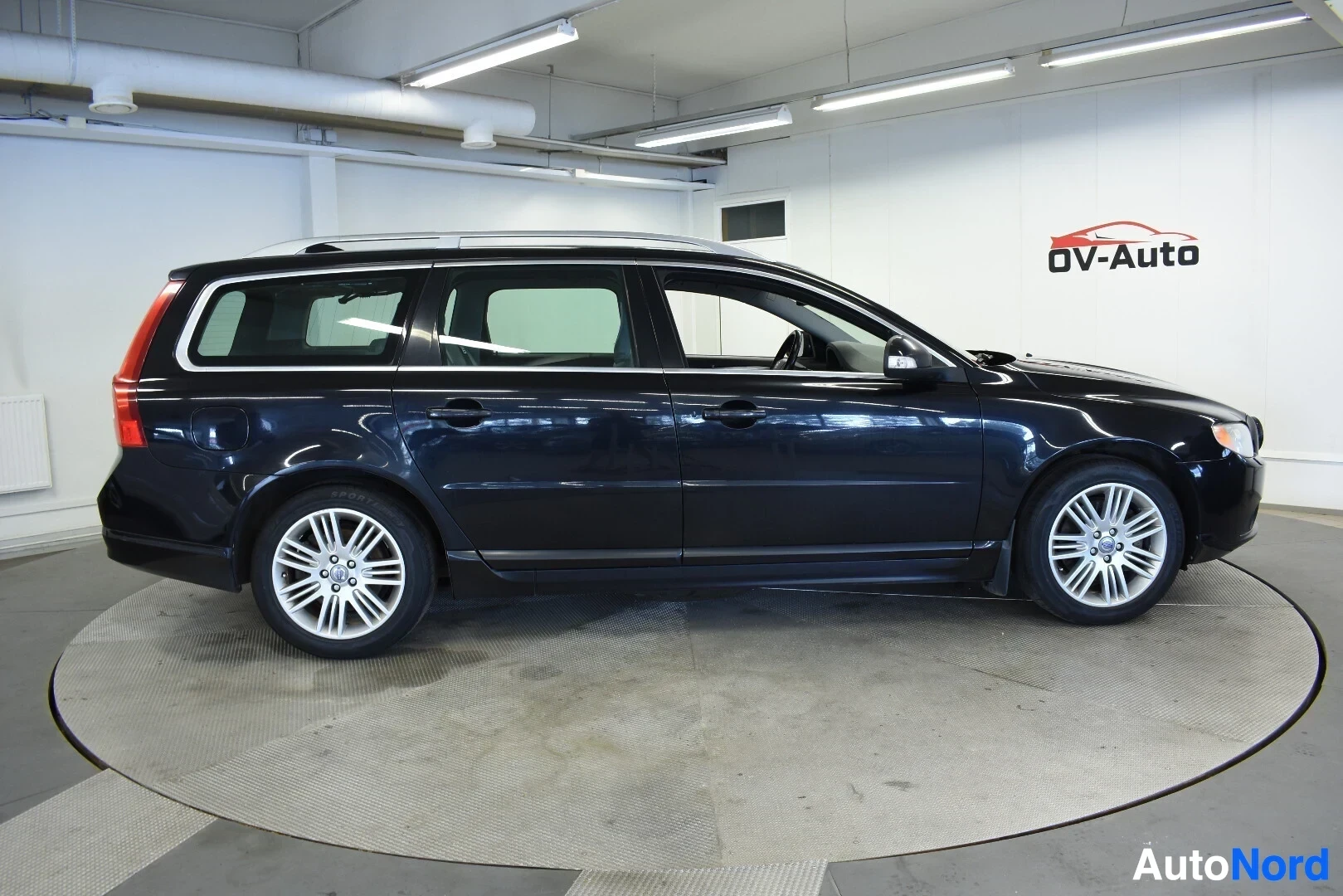 Sivulta: Musta Volvo V70 -farmariauto sivulta kuvattuna sisätiloissa. Autossa on kattokiskot.