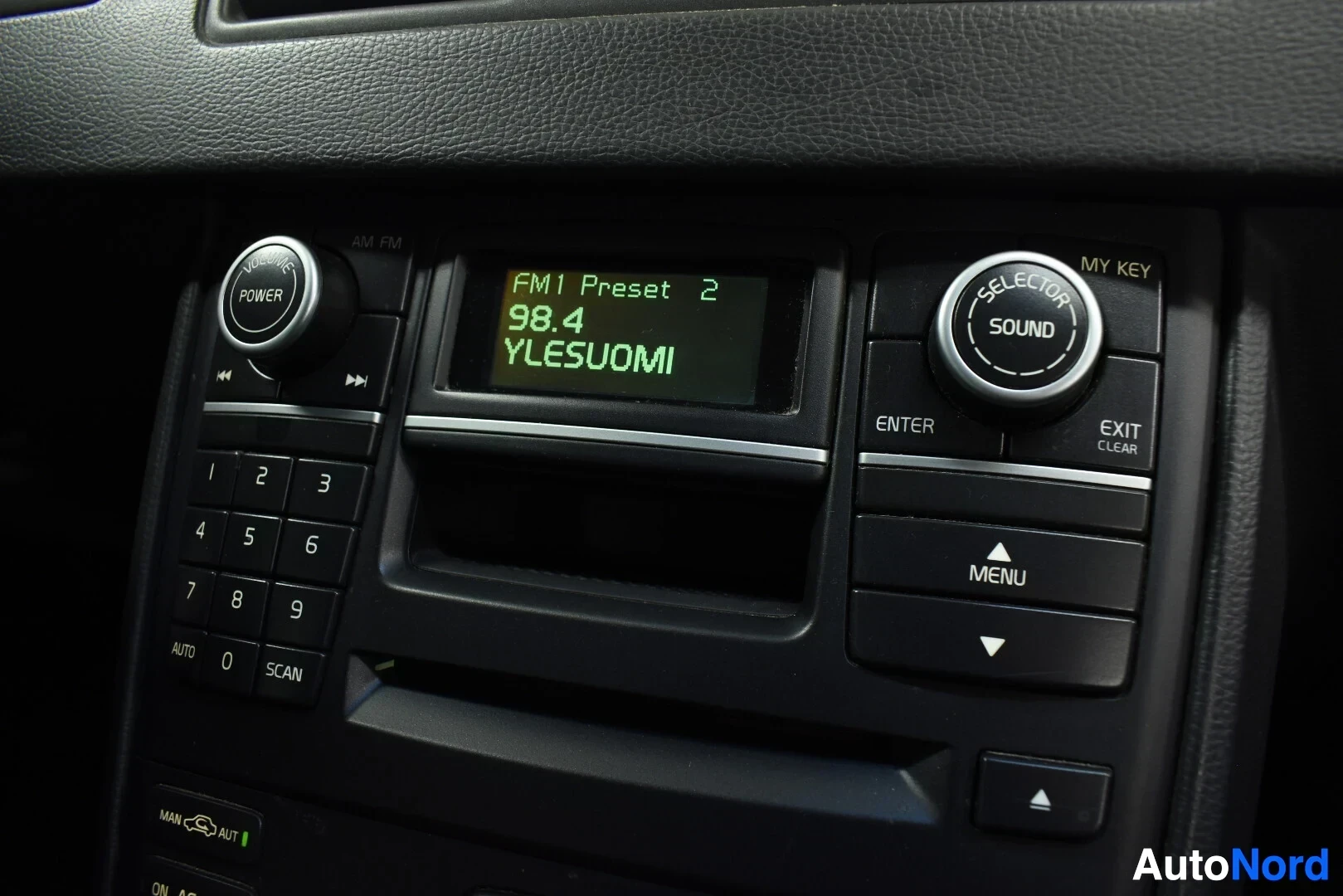 Yksityiskohta: Lähikuva auton radion ohjauspaneelista, jossa on näyttö, jossa lukee "FM1 Preset 2 98.4 YLESUOMI", ja numeronäppäimet 1-9 sekä virta- ja äänenvoimakkuussäätimet