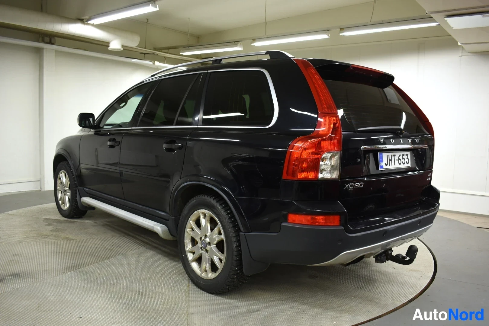 Takaviisto: Musta Volvo XC90:n takaoikea kulma, jossa on vetokoukku, kattotelineet ja hopeiset astinlaudat. Rekisterikilpi on ""