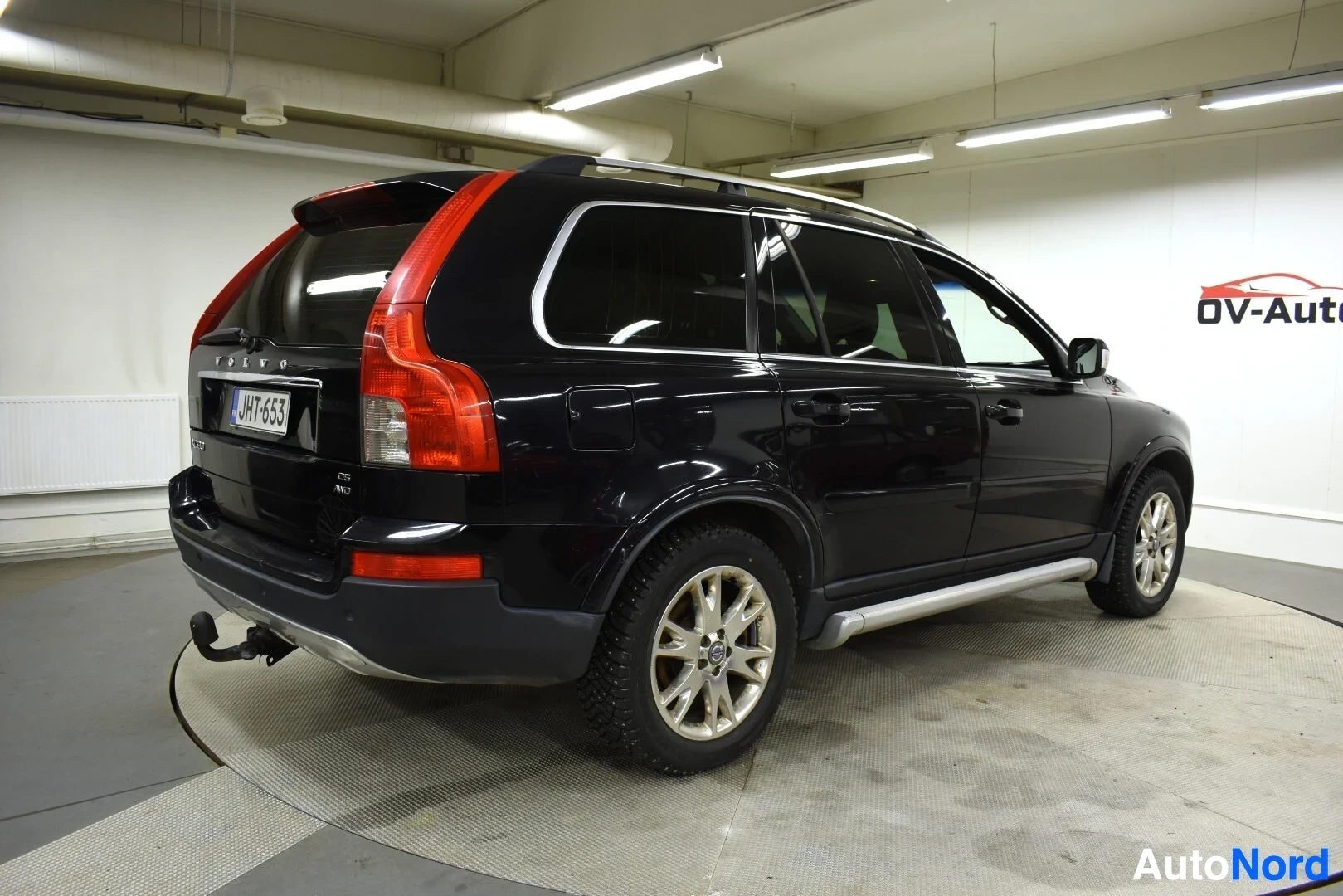 Takaviisto: Mustan Volvo XC90:n takavasen kulma, jossa on vetokoukku, kattotelineet ja hopeiset astinlaudat. Rekisterikilpi on ""