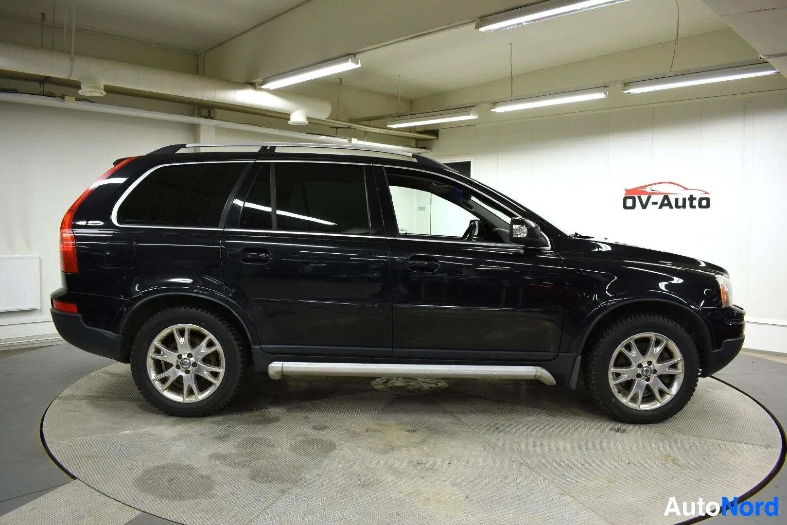 Sivulta: Mustan Volvo XC90:n vasen sivu, jossa on hopeiset kattotelineet ja astinlaudat. Autossa on viisipuolaiset kevytmetallivanteet.