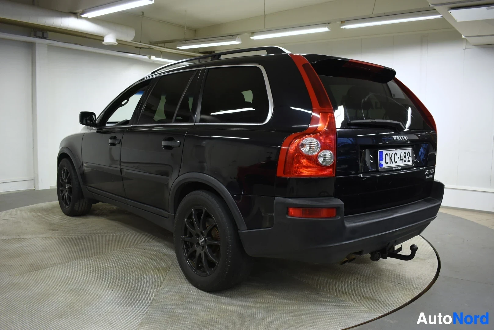Takaviisto: Musta Volvo XC90:n takaoikea kolmiokuvakulma pysäköitynä valkoisessa hallissa pyörivällä alustalla. Autossa on kattokaiteet ja vetokoukku.
