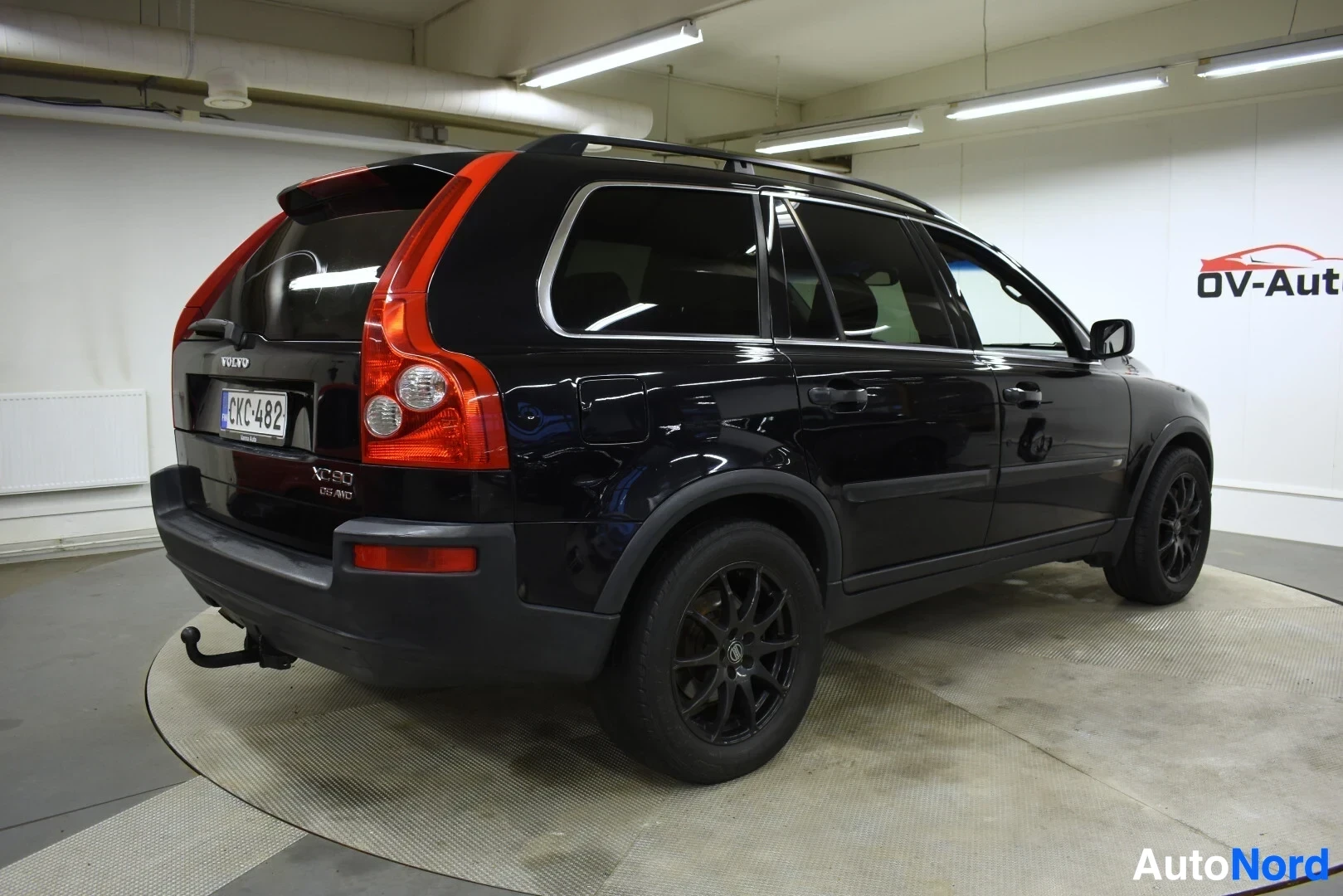 Takaviisto: Mustan Volvo XC90:n takavasen kolmiokuvakulma valkoisessa hallissa pyörivällä alustalla. Autossa on kattokaiteet ja vetokoukku.