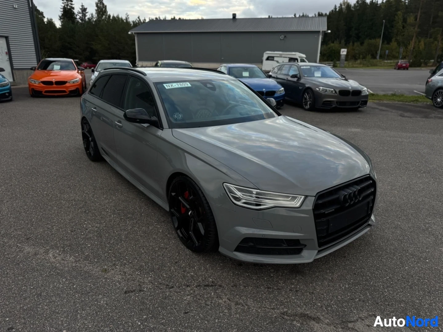Etuviisto, nokka vasemmalle: Harmaa farmariauto, jossa on mustat monipuolaiset kevytmetallivanteet ja punaiset jarrusatulat, pysäköity asvalttipinnalle muita autoja ja rakennuksia
