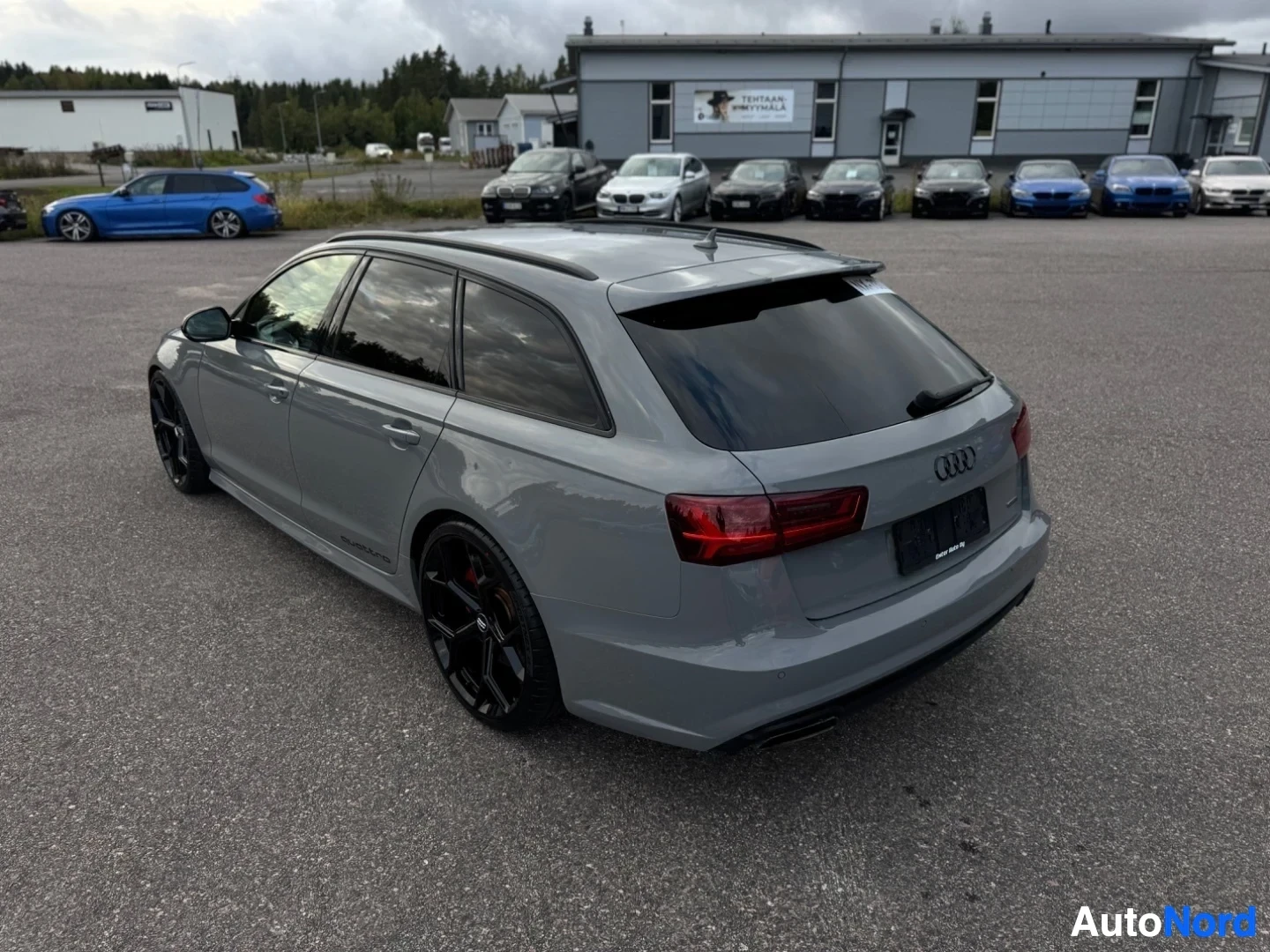 Takaviisto: Harmaa farmariauto, jossa on mustat kevytmetallivanteet ja punaiset jarrusatulat, pysäköity ulos muiden autojen ja rakennusten eteen. Auton takaluukun oikeassa